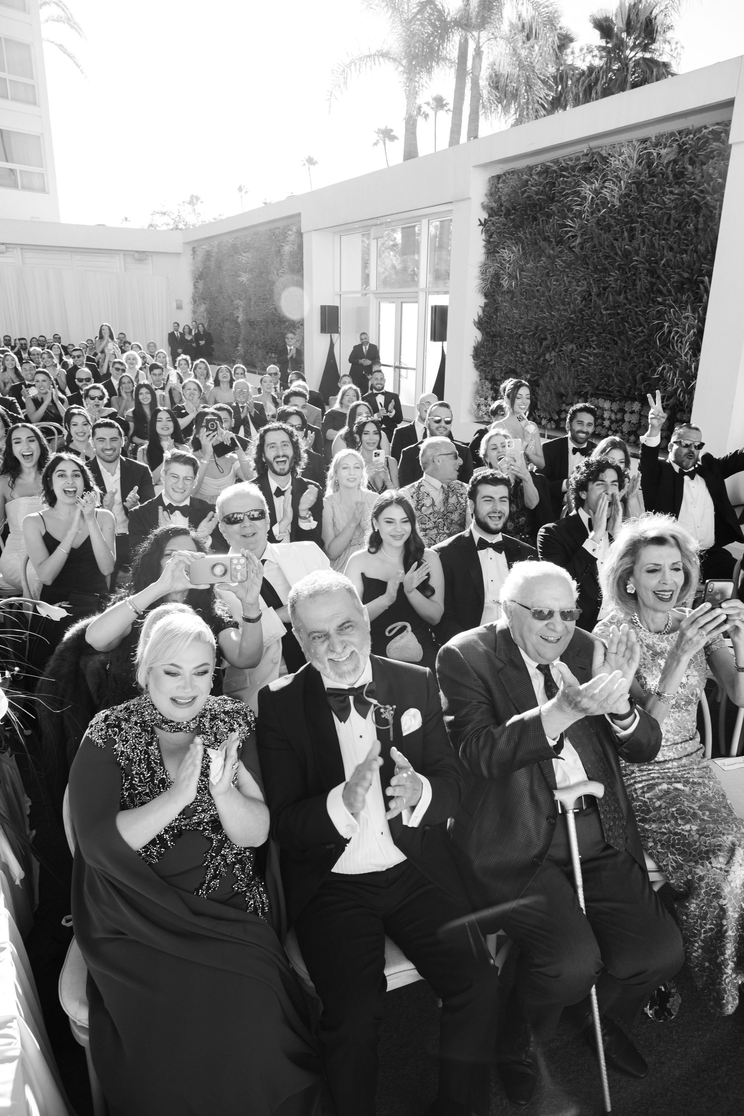 Black and white photo of elegantly dressed guests clapping and smiling at an outdoor formal event or awards ceremony.