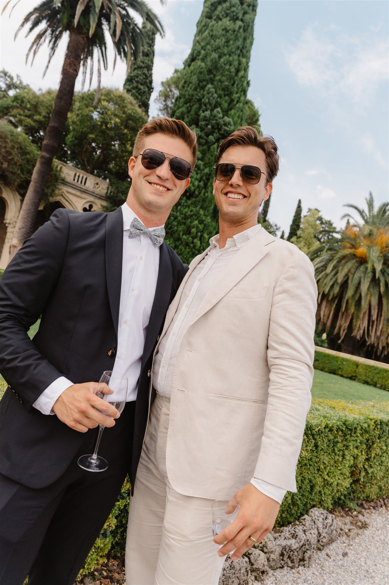 Wedding guests in suits and sunglasses enjoying drinks in a Mediterranean garden setting.