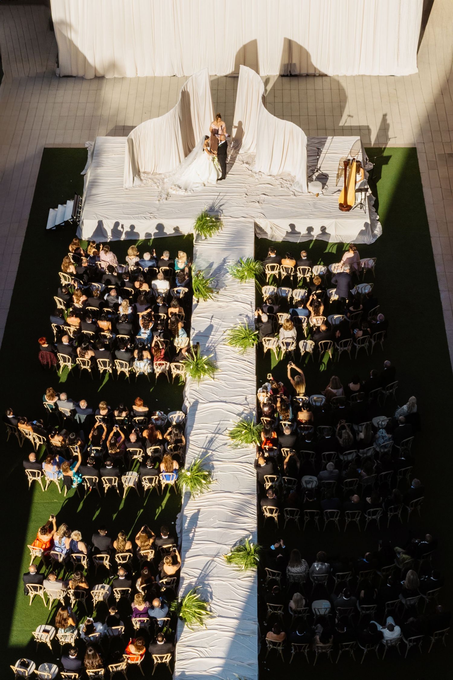 Aerial view of an outdoor wedding ceremony with white aisle, decorated altar, and rows of seated guests on green lawn.