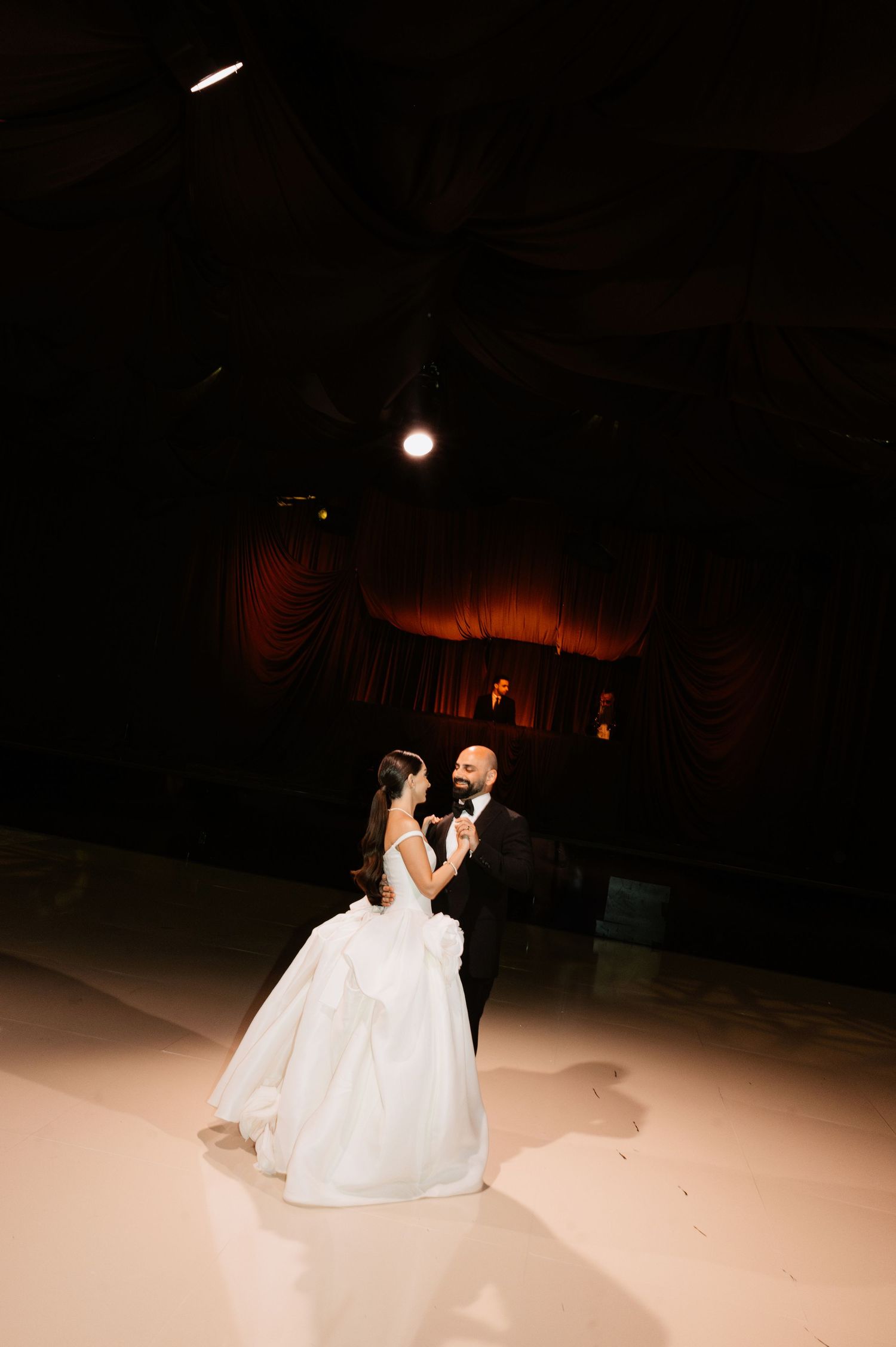 Bride and groom share their first dance on a lit dance floor in an elegant dark venue with red draped curtains.