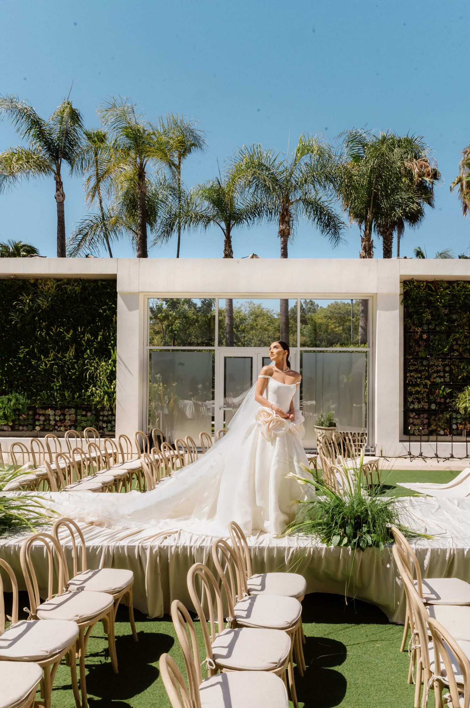 Bride in a flowing white gown stands at an outdoor wedding ceremony venue with palm trees and rattan chairs.