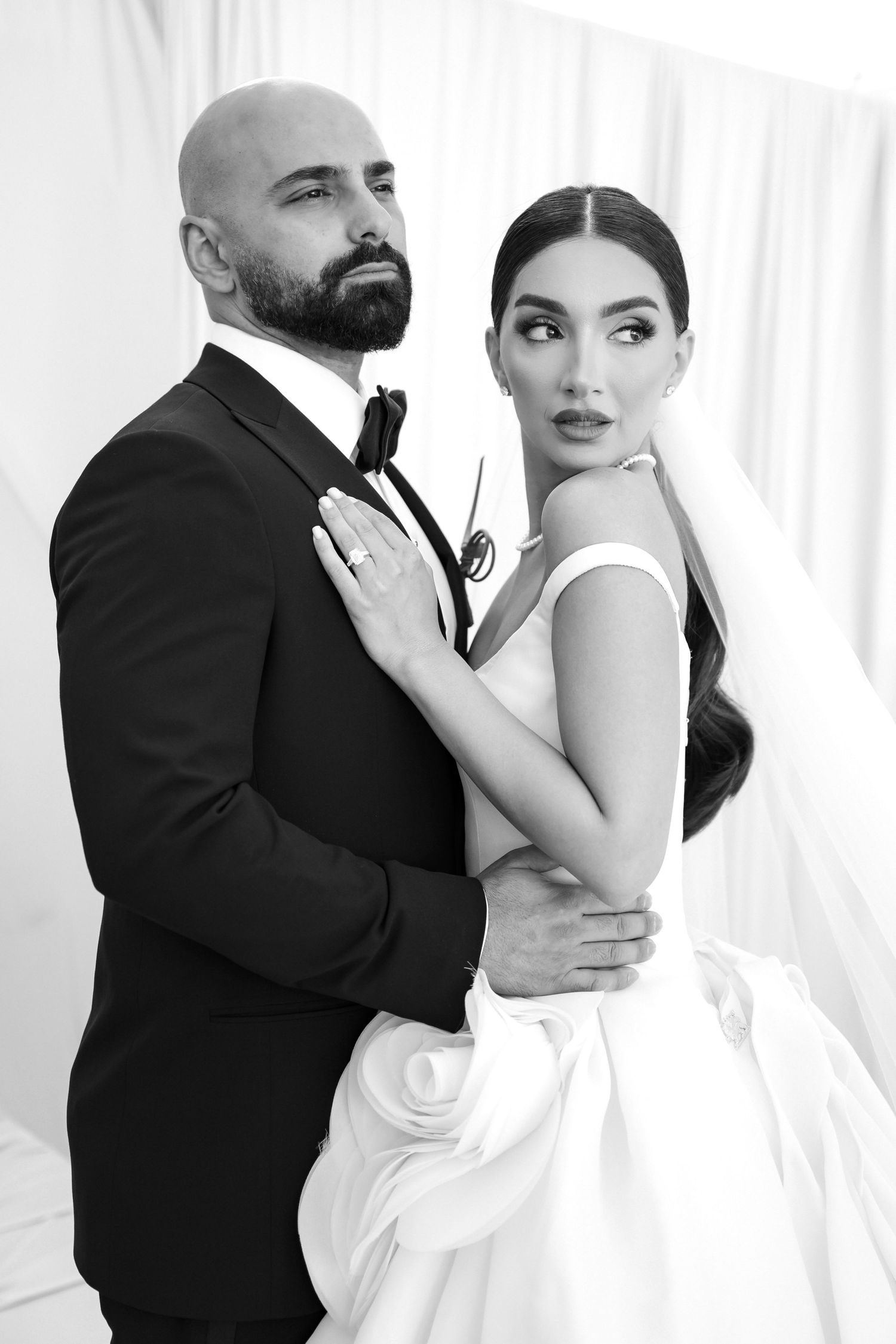 Black and white wedding portrait of elegant bride in gown embracing bald groom in tuxedo.