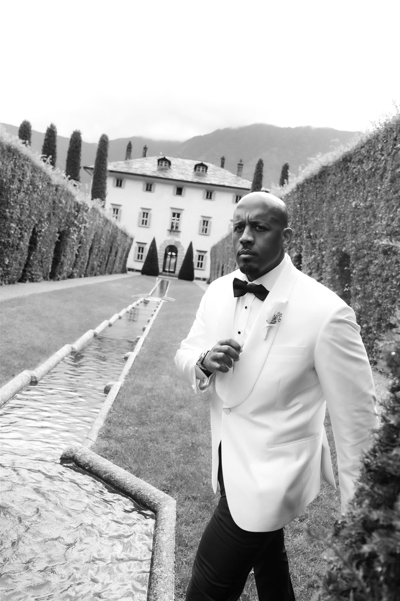 Person in white tuxedo posing dramatically in formal garden with stone pathway.