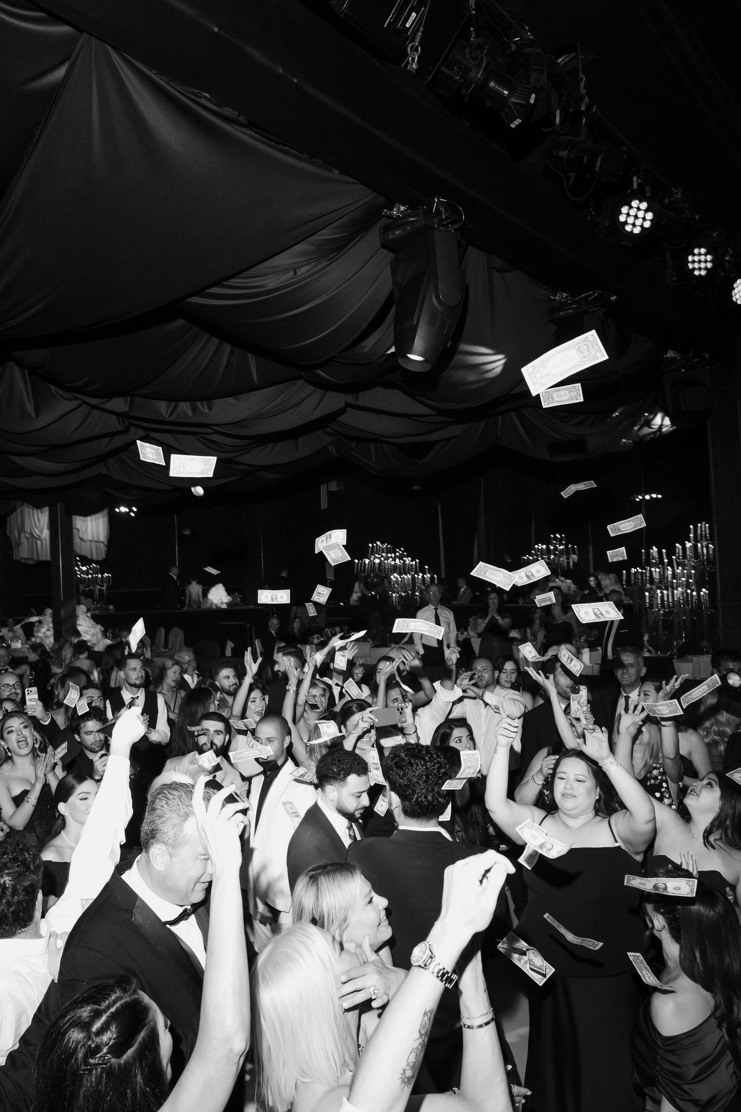 Black and white photo of a lively formal event crowd waving papers in the air under stage lights.