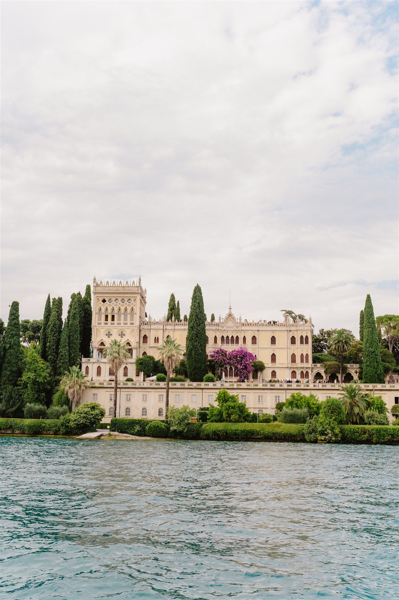 Historic Italian villa with cypress trees and ornate architecture sits majestically along Lake Garda's waterfront.