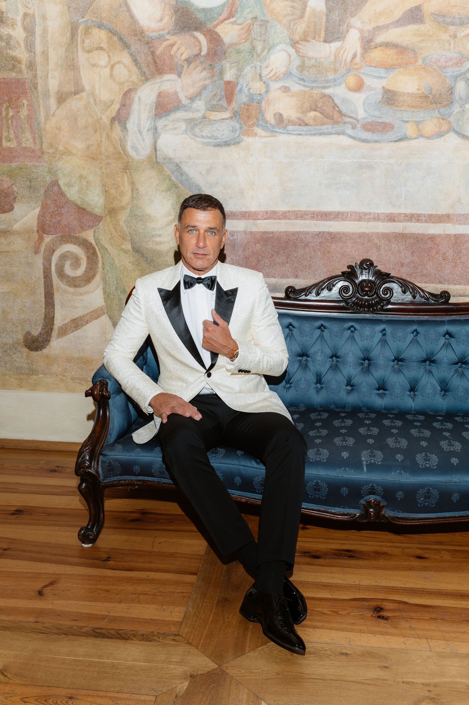 Person in tuxedo lounging on blue velvet sofa against decorated wall.