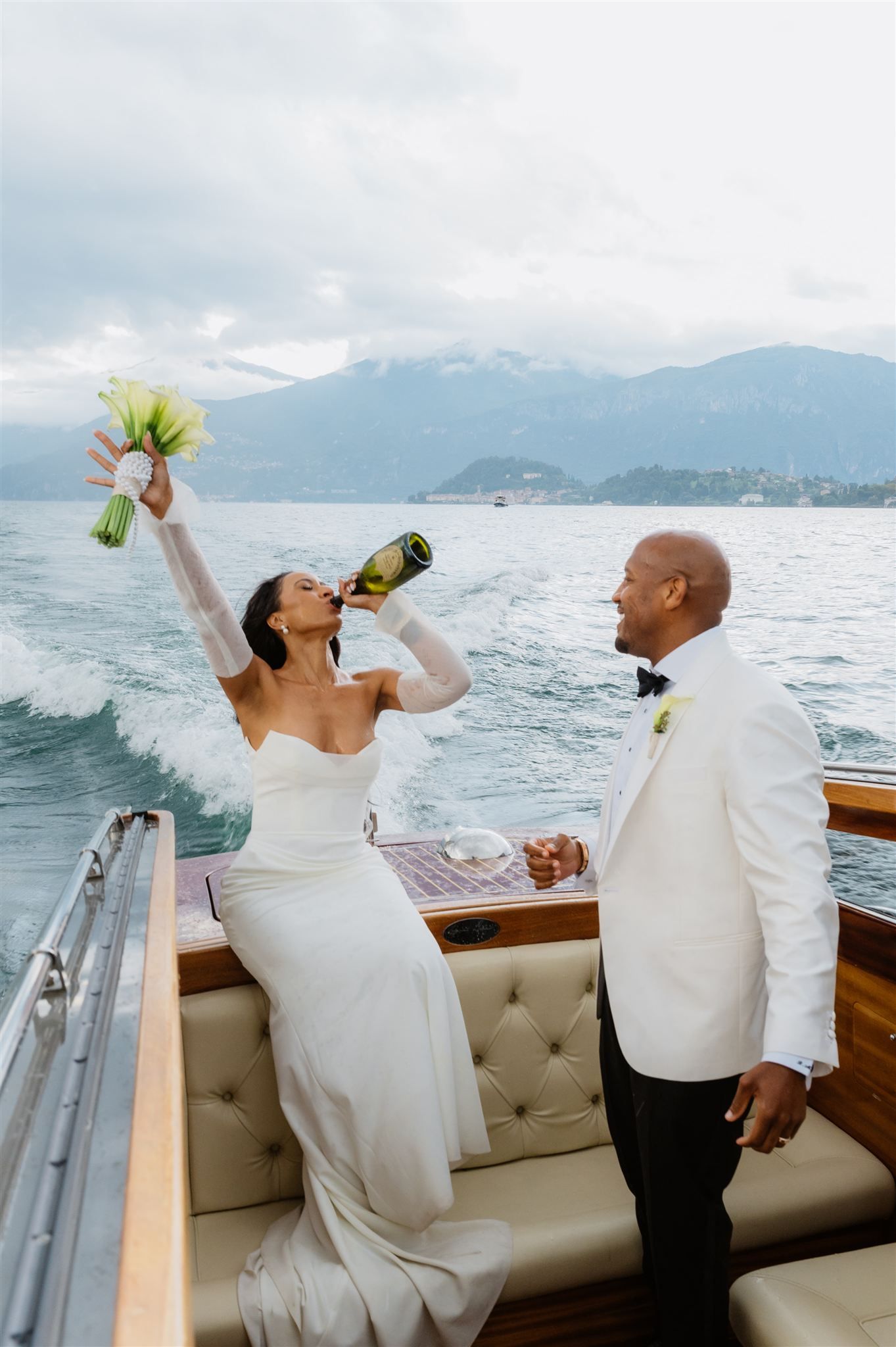 Couple in white formal wear toasting with champagne on a wooden boat on Lake Como
