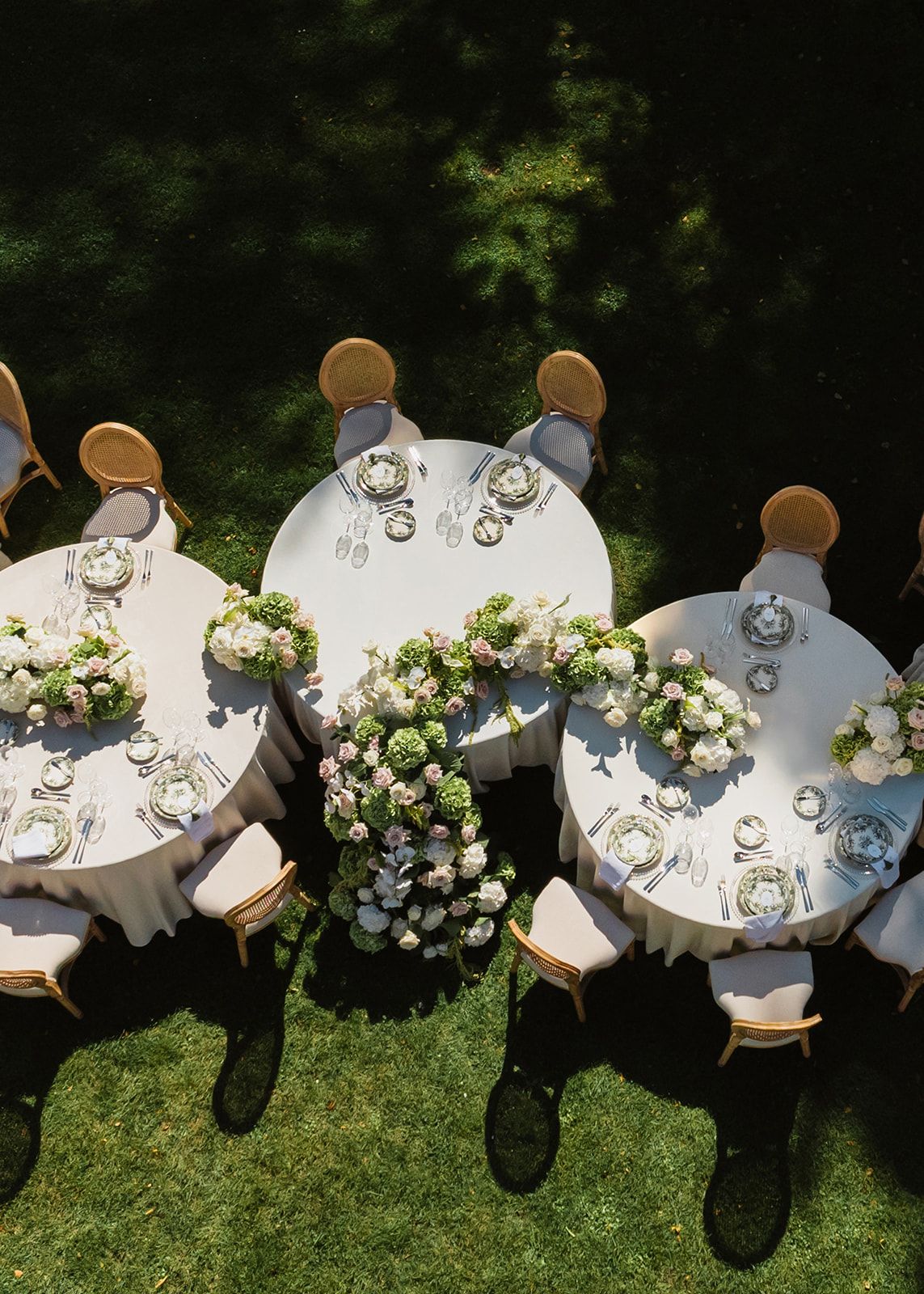 Elegant outdoor wedding reception setup with round tables and white linens decorated with floral centerpieces on a grass lawn.