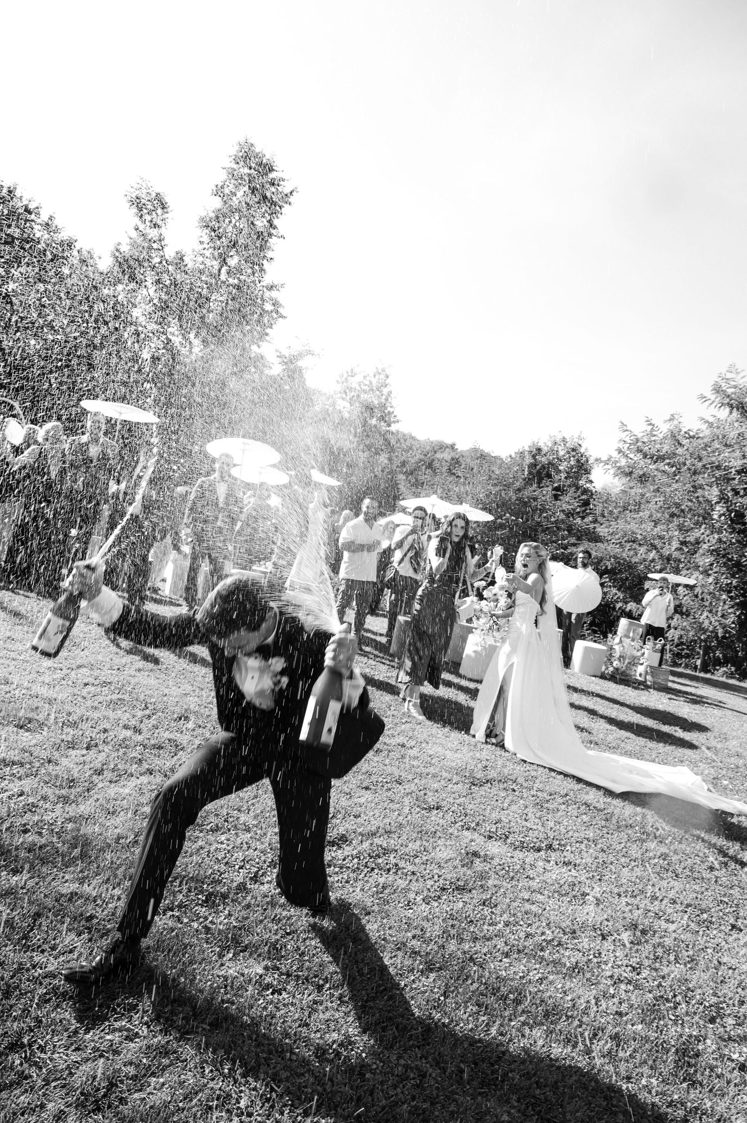 Black and white photo of guests celebrating at an outdoor wedding reception with champagne spray. Candid