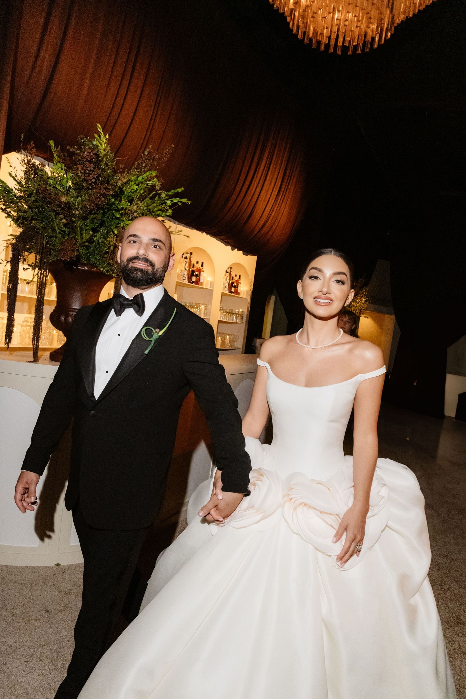 Bride in white off-shoulder gown and groom in black tuxedo hold hands smiling at elegant wedding reception.