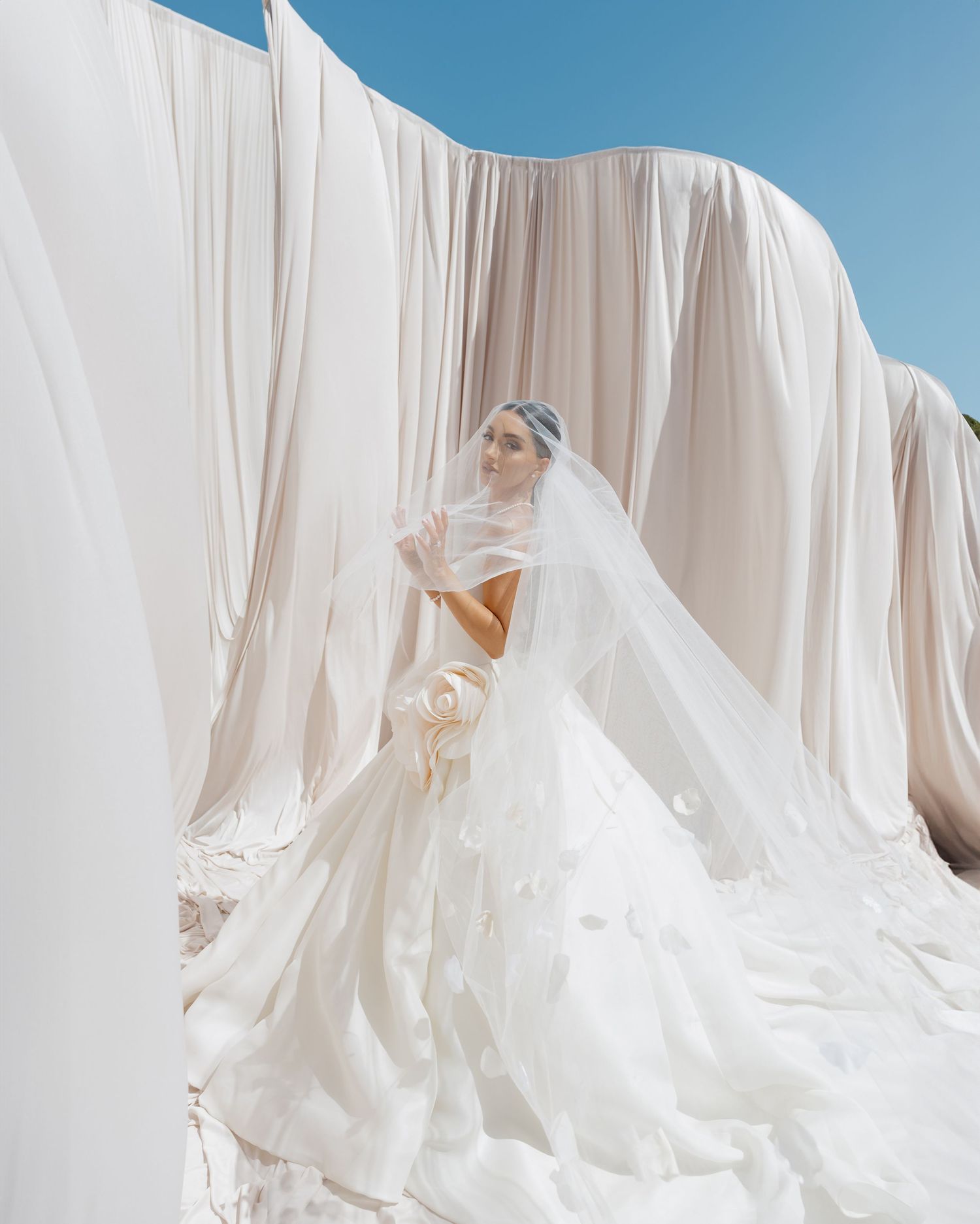 Bride in a stunning white ball gown with veil poses gracefully among flowing ivory drapes under a blue sky.