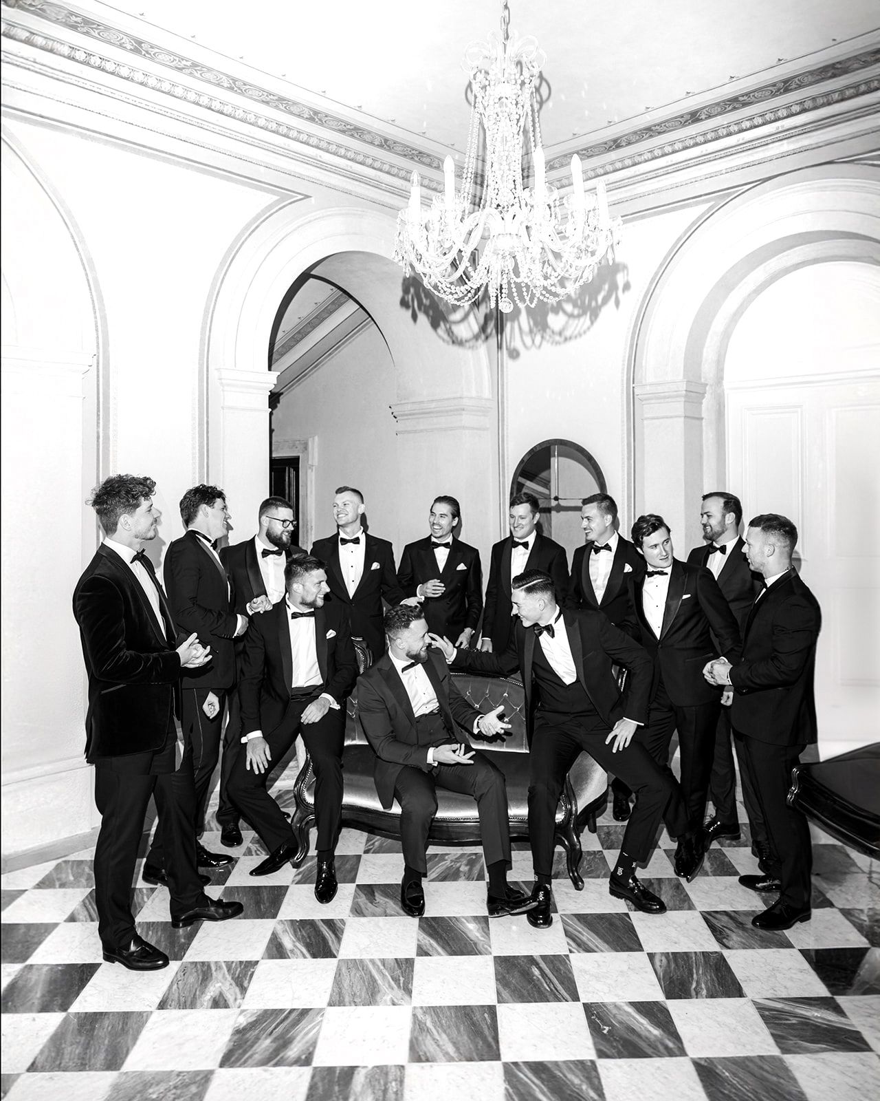 Groomsmen in black suits share laughter and camaraderie in an elegant room with chandeliers and checkered floors.