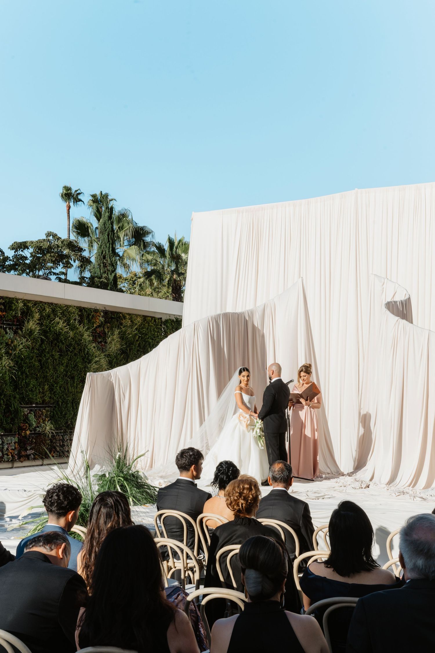 Outdoor wedding ceremony with bride and groom at draped white backdrop altar, guests seated in wooden chairs under blue sky.