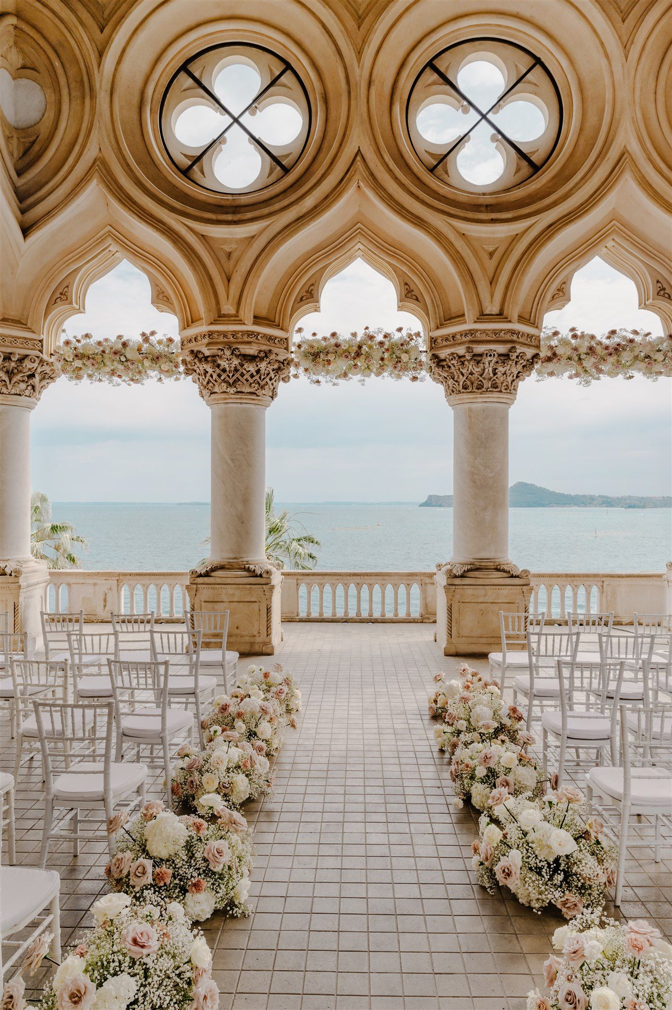 Ornate Gothic arches frame a lakeside wedding ceremony setup with white floral arrangements.