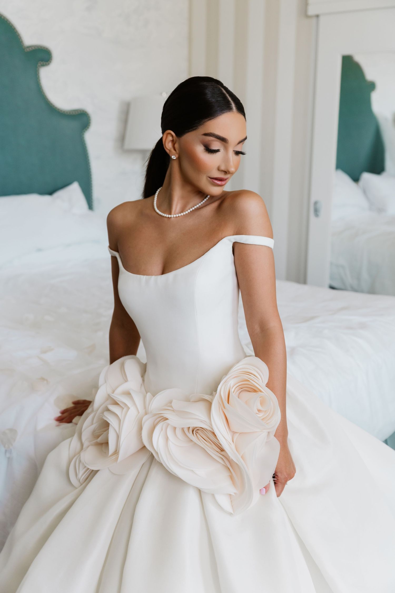 Bride in off-shoulder white ball gown with floral detail and pearl necklace sits elegantly on bed in bridal suite.