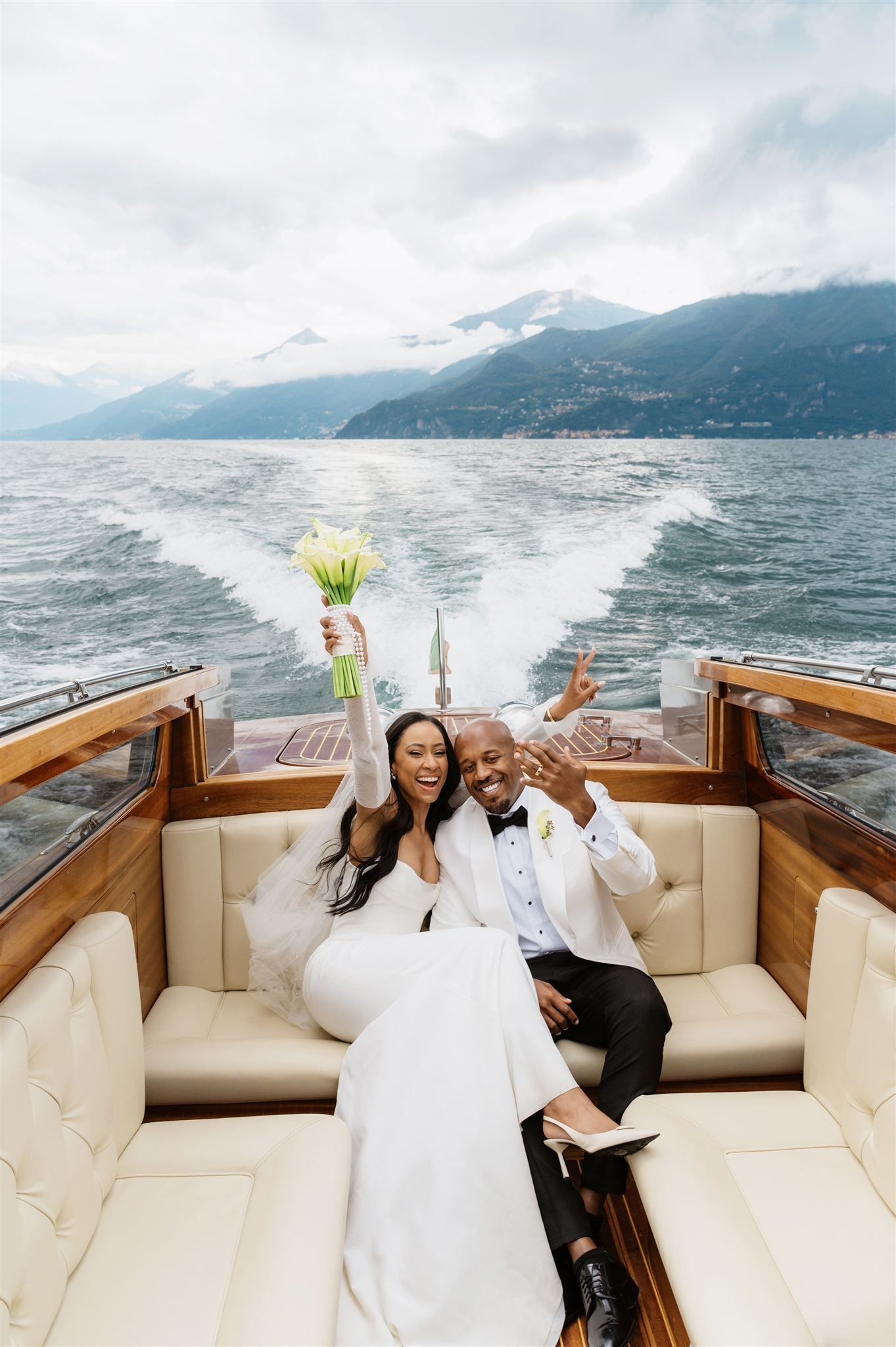 Happy couple in formal wear celebrating with drinks on a boat against mountain backdrop on Lake Como