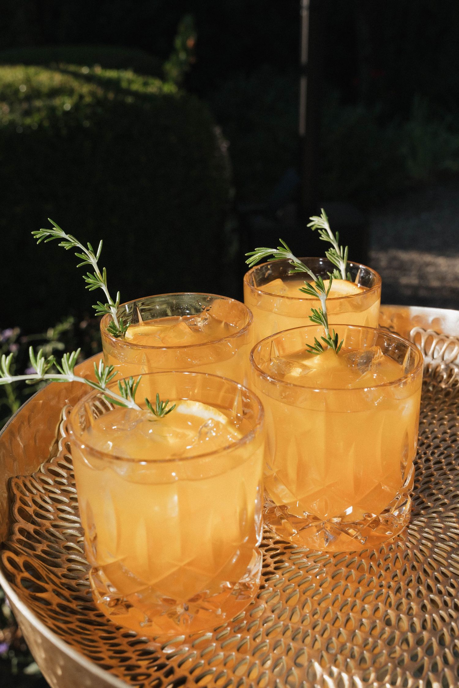 Four orange cocktails garnished with fresh rosemary sprigs served on a textured tray.