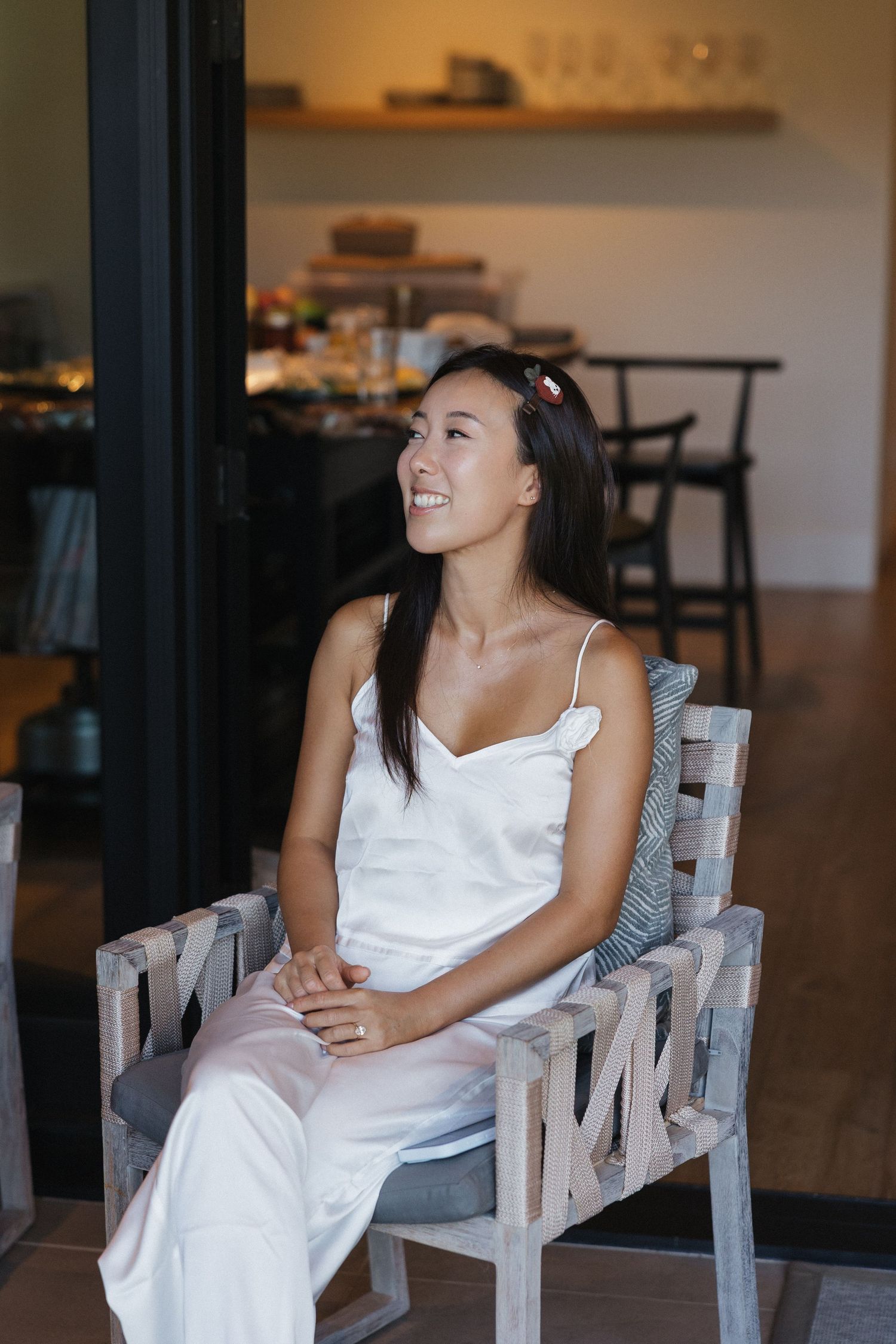 A person in a white dress sits thoughtfully on a wooden chair in an indoor setting with warm lighting.
