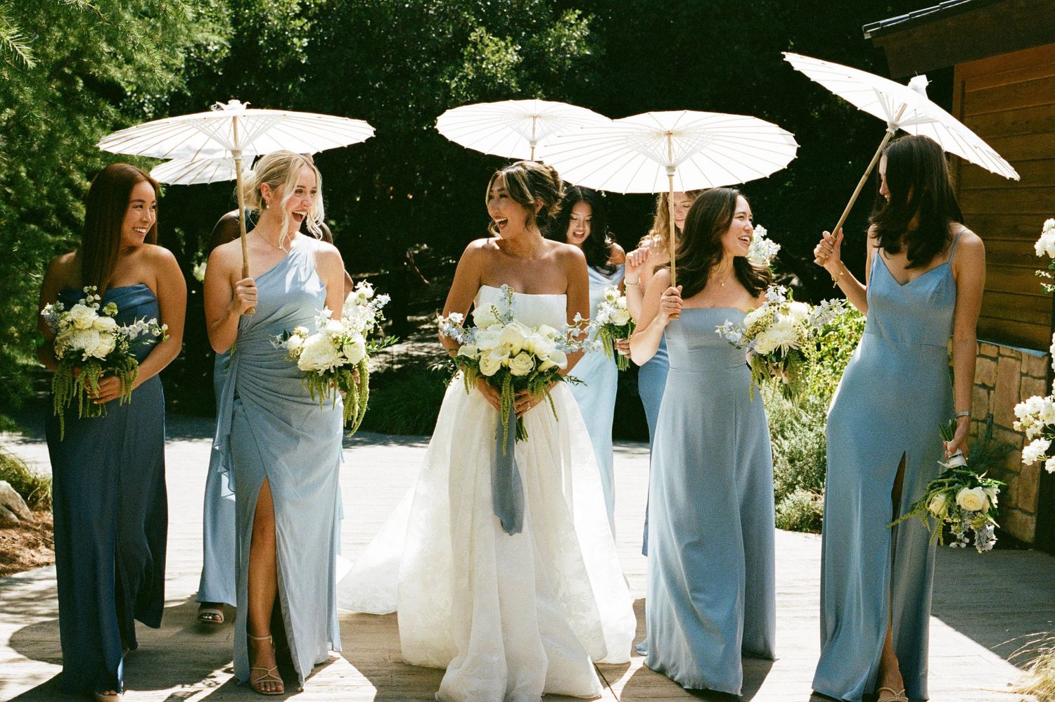 Bridesmaids in light blue dresses holding white parasols and bouquets pose together outdoors.