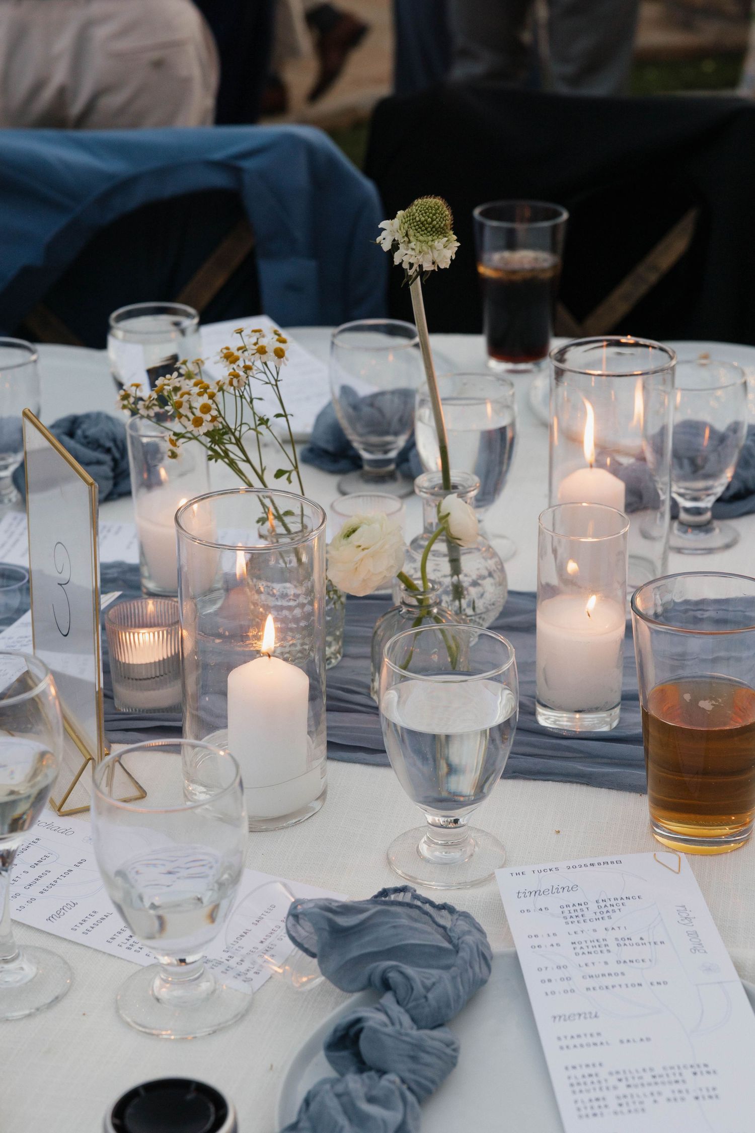 An elegant table setting with white candles in glass holders, simple floral accents, and table menus on a grey tablecloth.