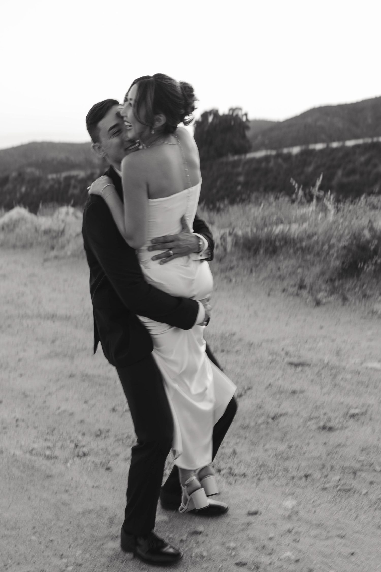 Black and white candid shot of newlyweds embracing on dirt path with hills in background.