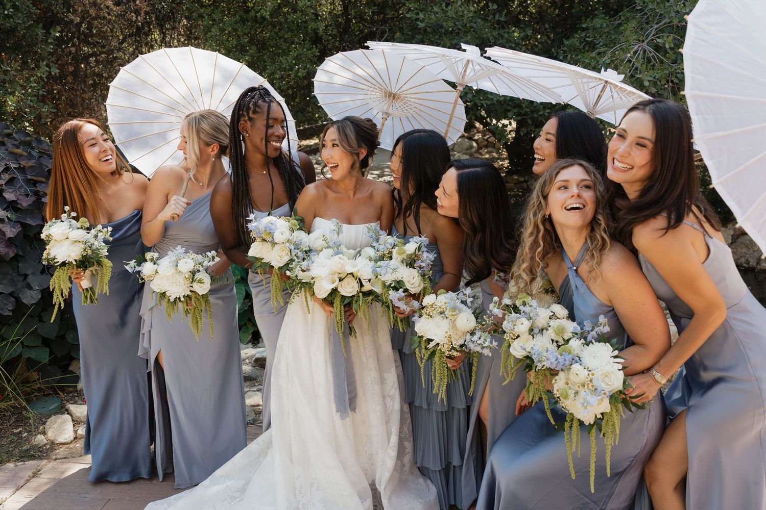 Wedding party with white parasols and bouquets pose together in pale blue bridesmaid dresses.