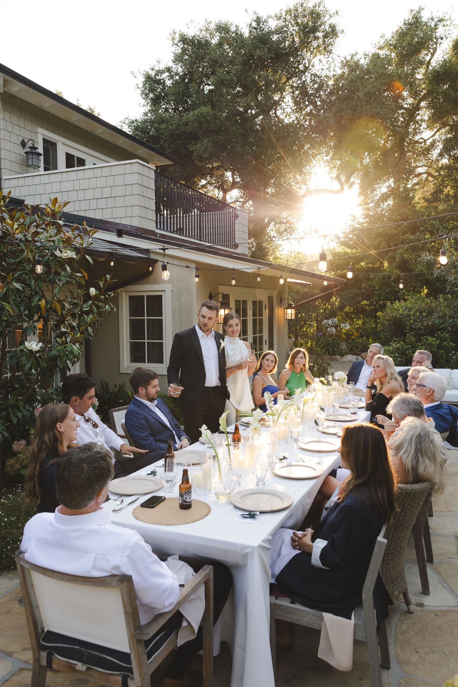 Outdoor dinner party at sunset with guests seated at a long white-clothed table on a patio under string lights.