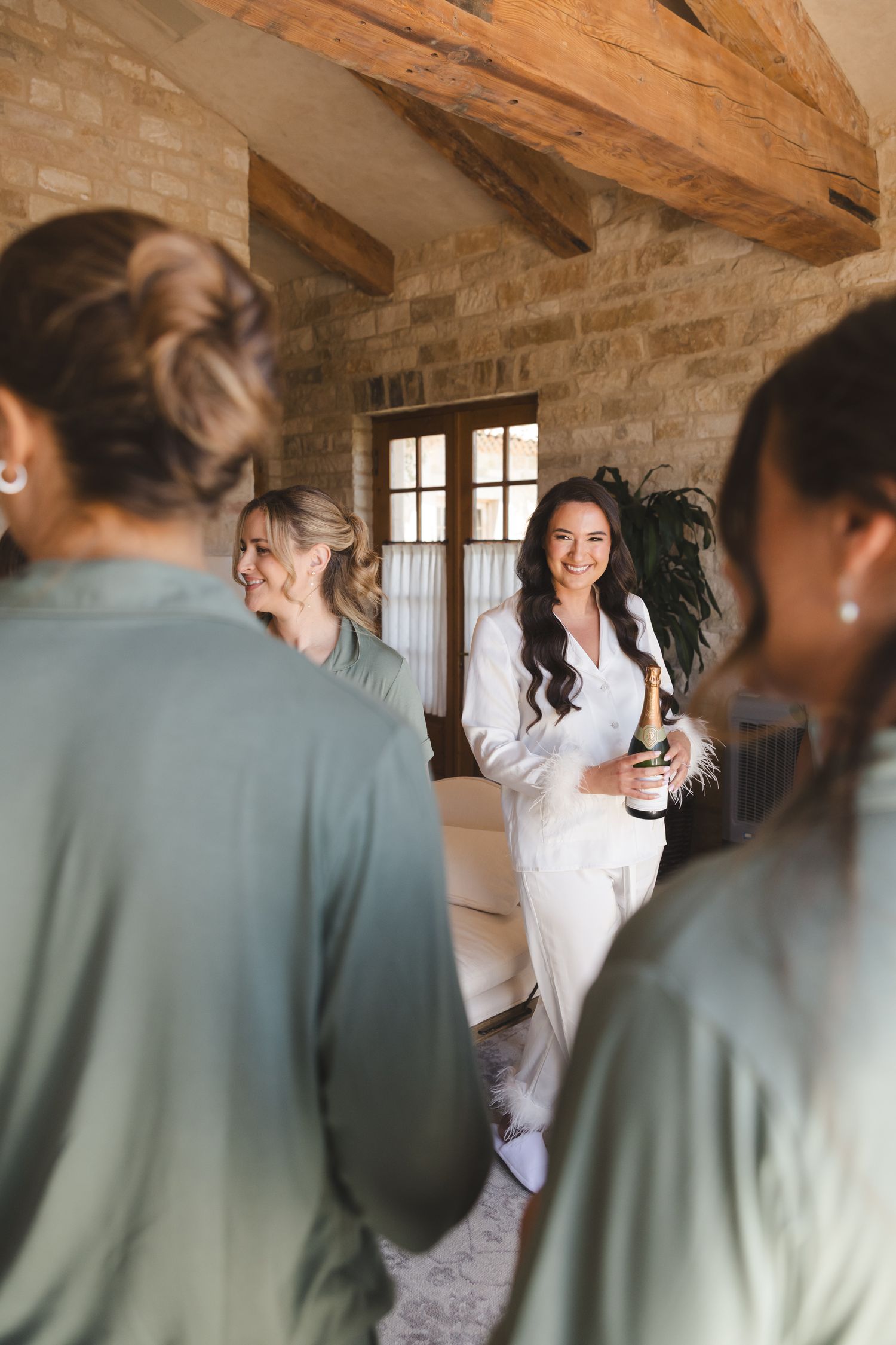 People gather in rustic room with exposed wooden beams and stone walls as person in white dress stands holding bottle.