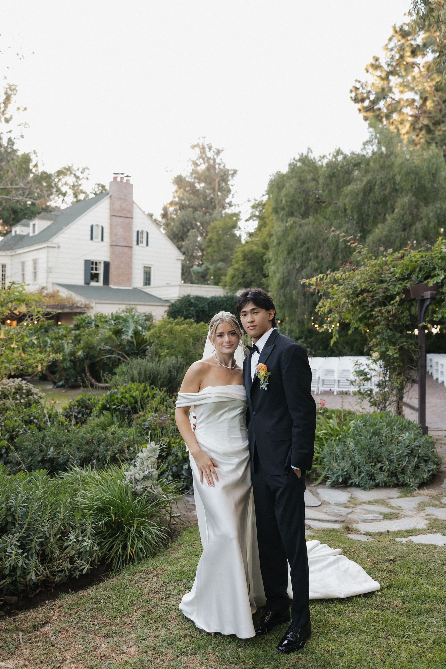 A couple poses for a wedding portrait in front of a white house with lush garden landscaping.