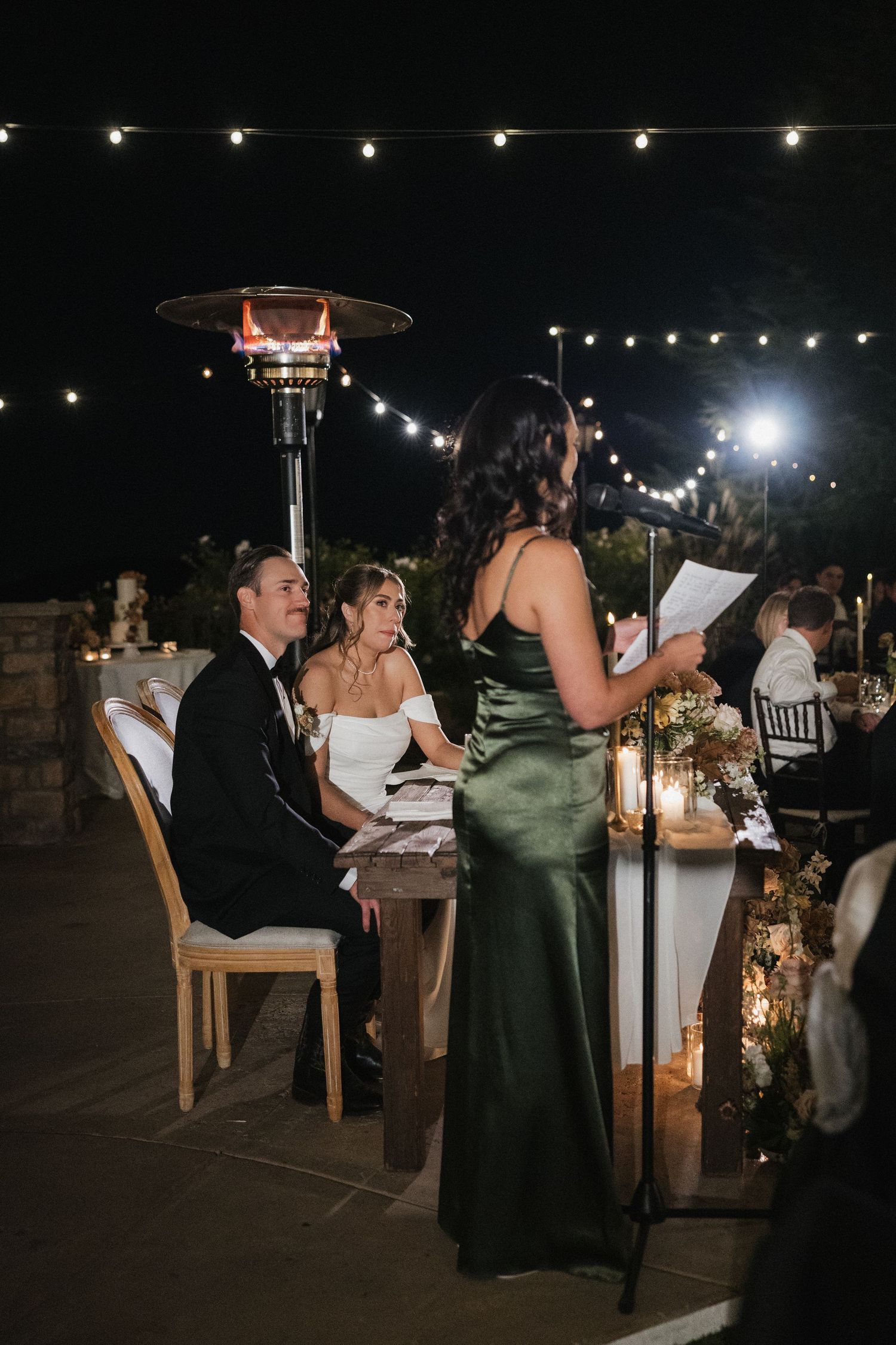 Wedding reception guests dine at tables under string lights at an elegant outdoor evening celebration.