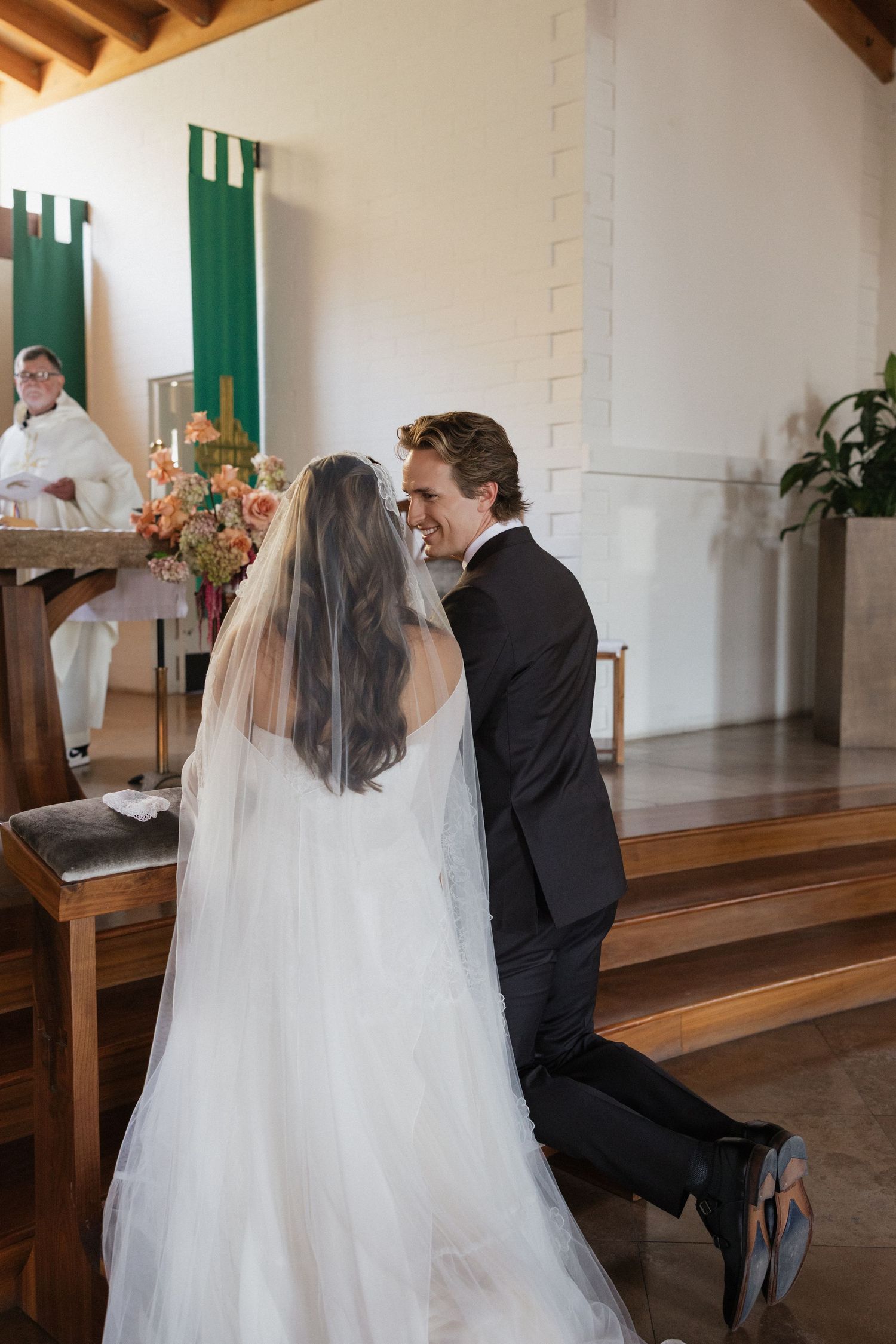 Bride and groom kneeling at altar during Catholic wedding ceremony with priest officiating.