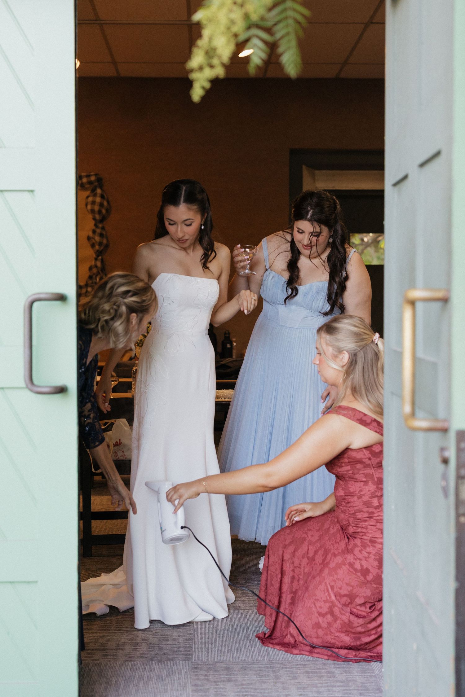 Bride in white strapless gown being helped by bridesmaids in blue and a woman in red dress in a barn doorway.