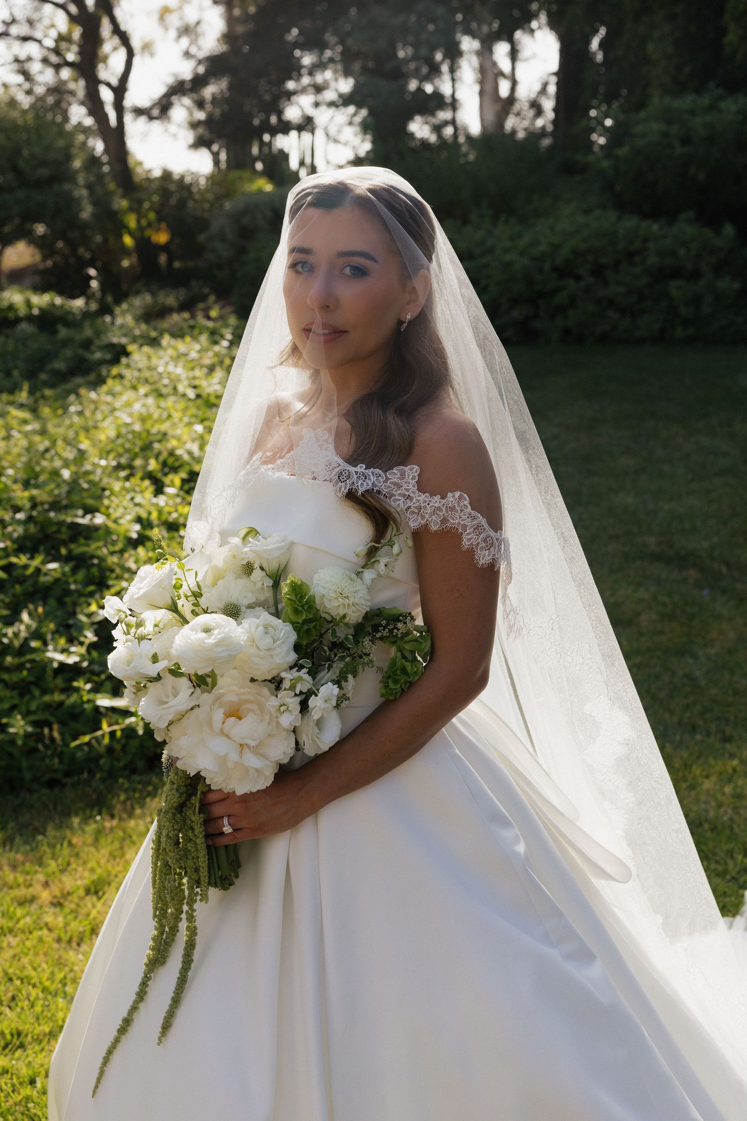 Bride in white off-shoulder gown and veil holds white peony bouquet outdoors in sunlit garden.