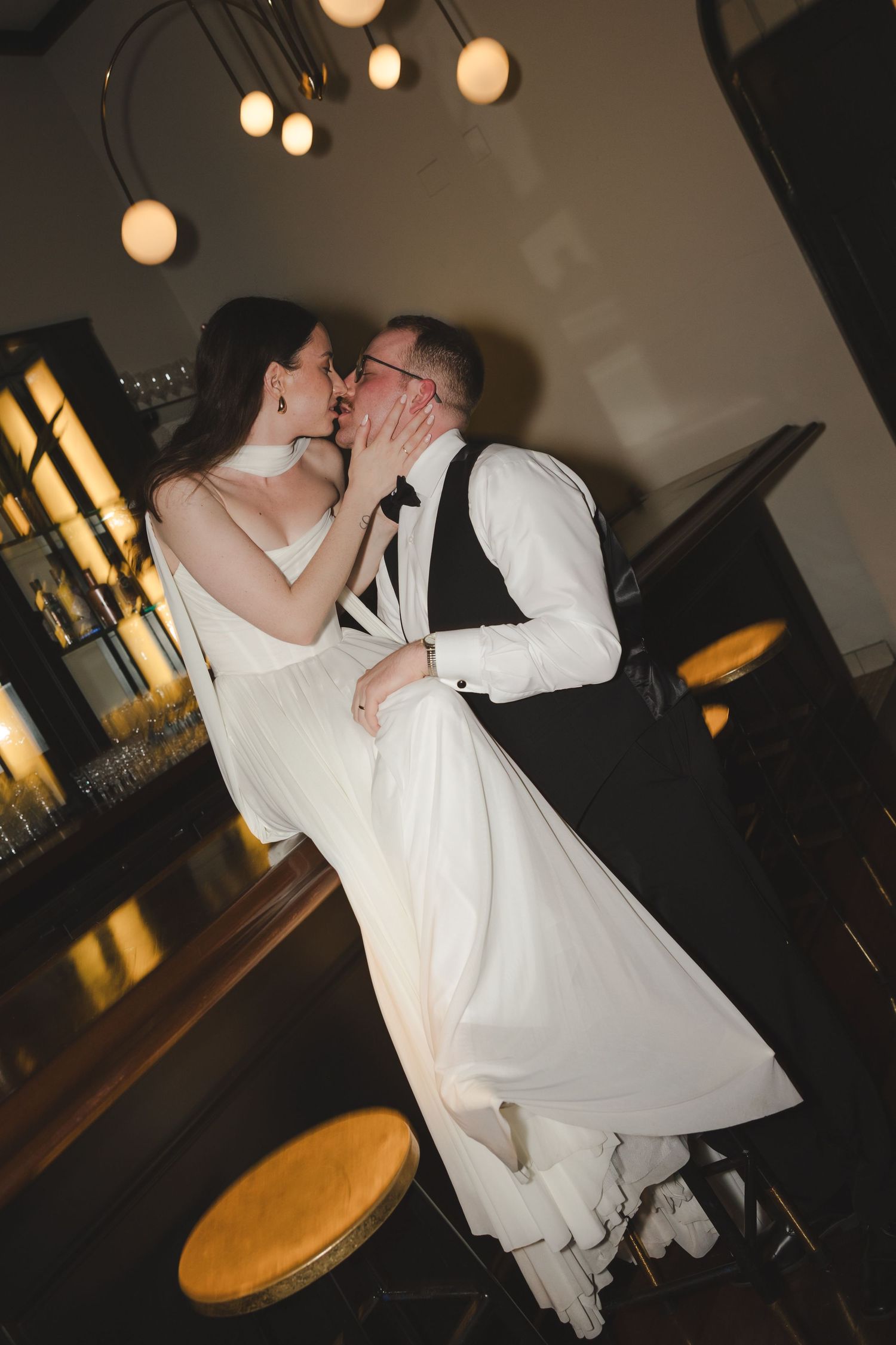 A couple shares a romantic kiss in a dimly lit venue with glowing pendant lights above during their wedding celebration.