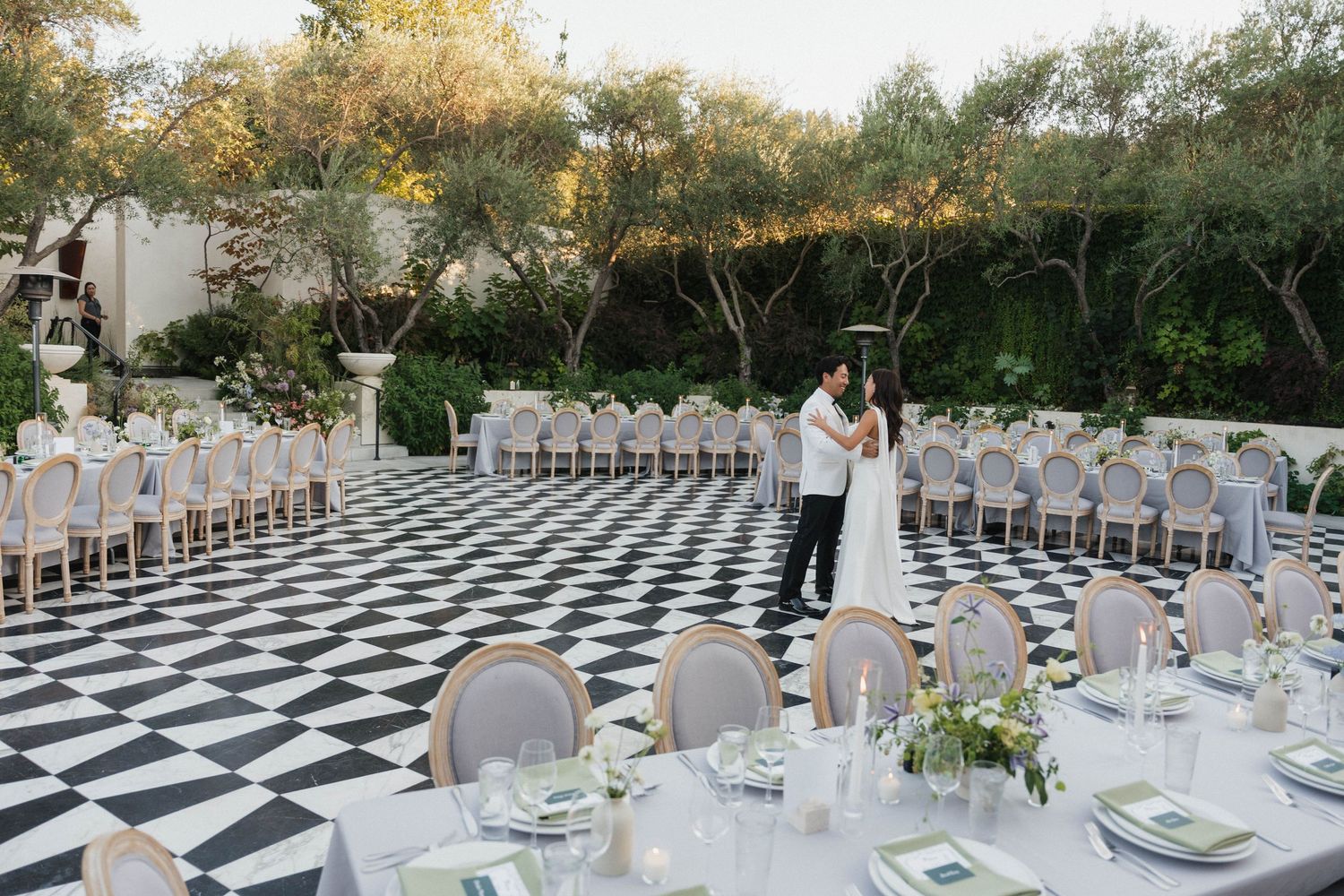 Elegant wedding reception setup with black and white checkered dance floor and white chairs arranged under trees at sunset.