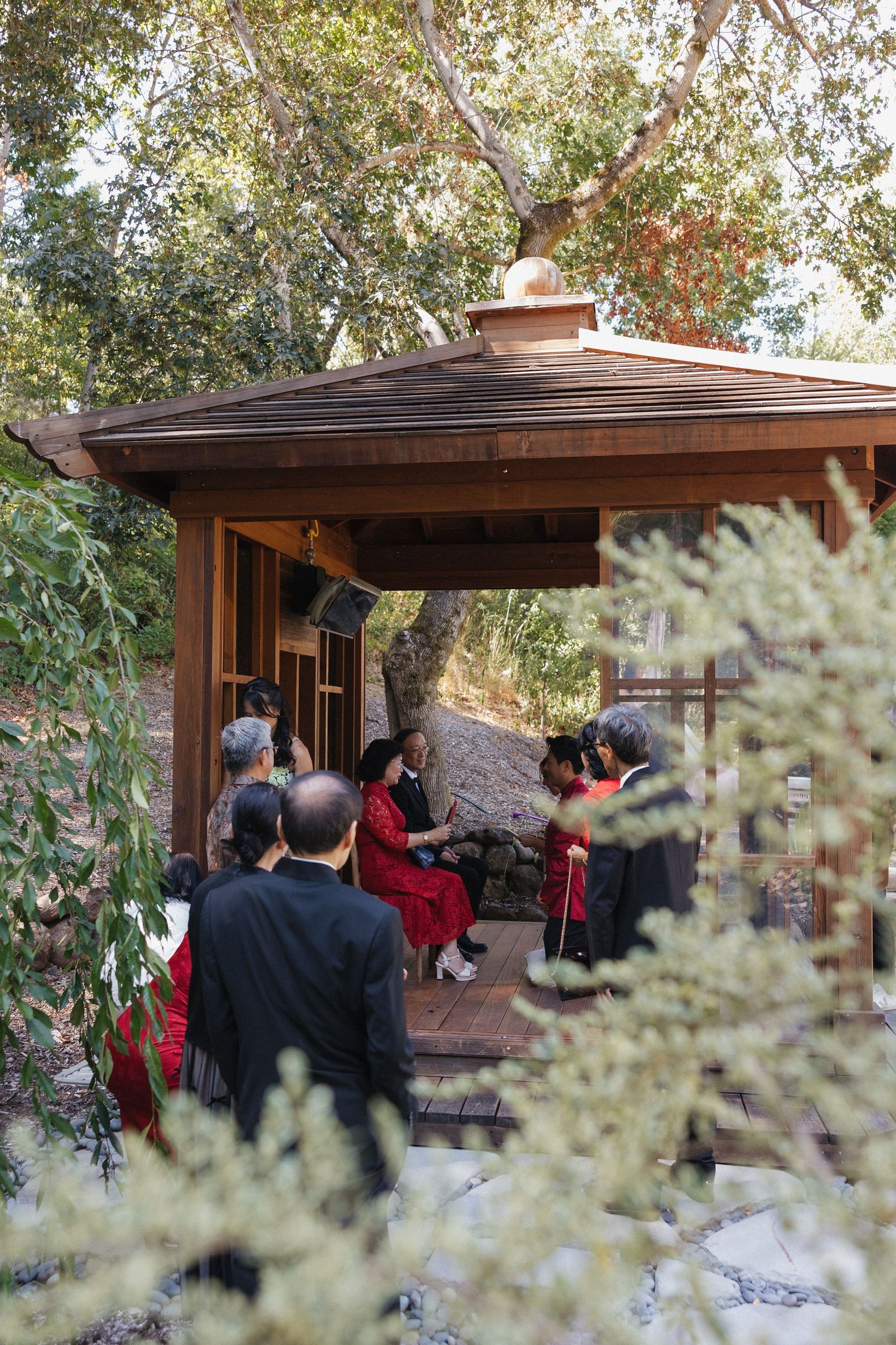 Wedding guests gather under a wooden gazebo surrounded by lush greenery during an outdoor ceremony.