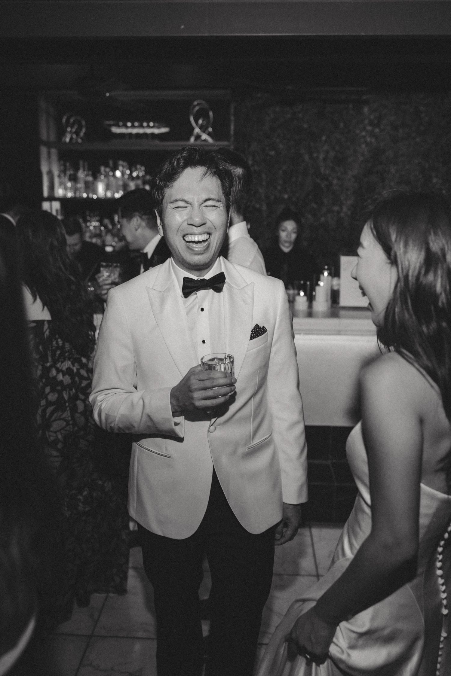 Wedding reception guests in formal attire enjoying drinks and conversation in dark venue.
