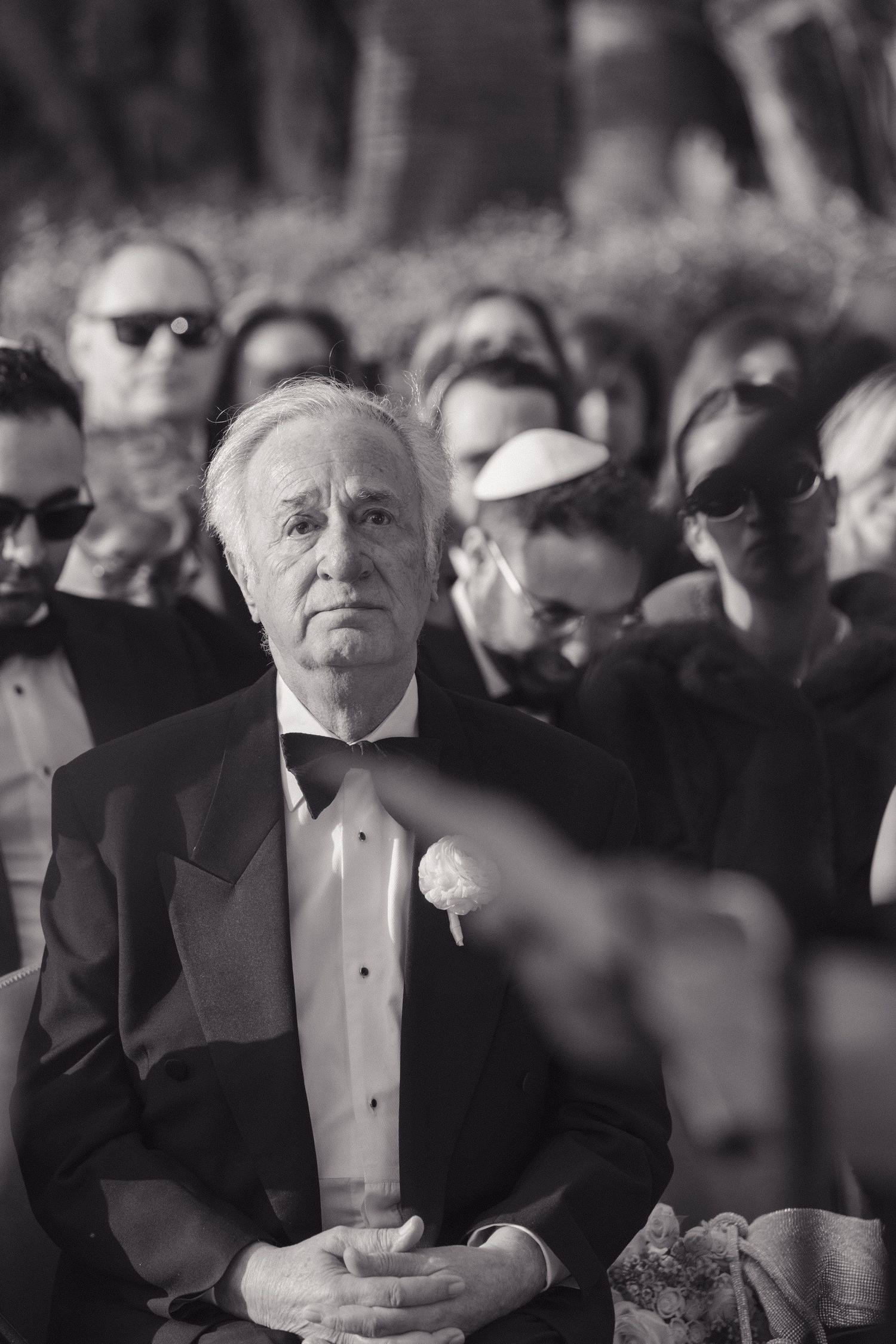 An elderly man in formal black tuxedo and bow tie sits solemnly among a crowd at an elegant event.