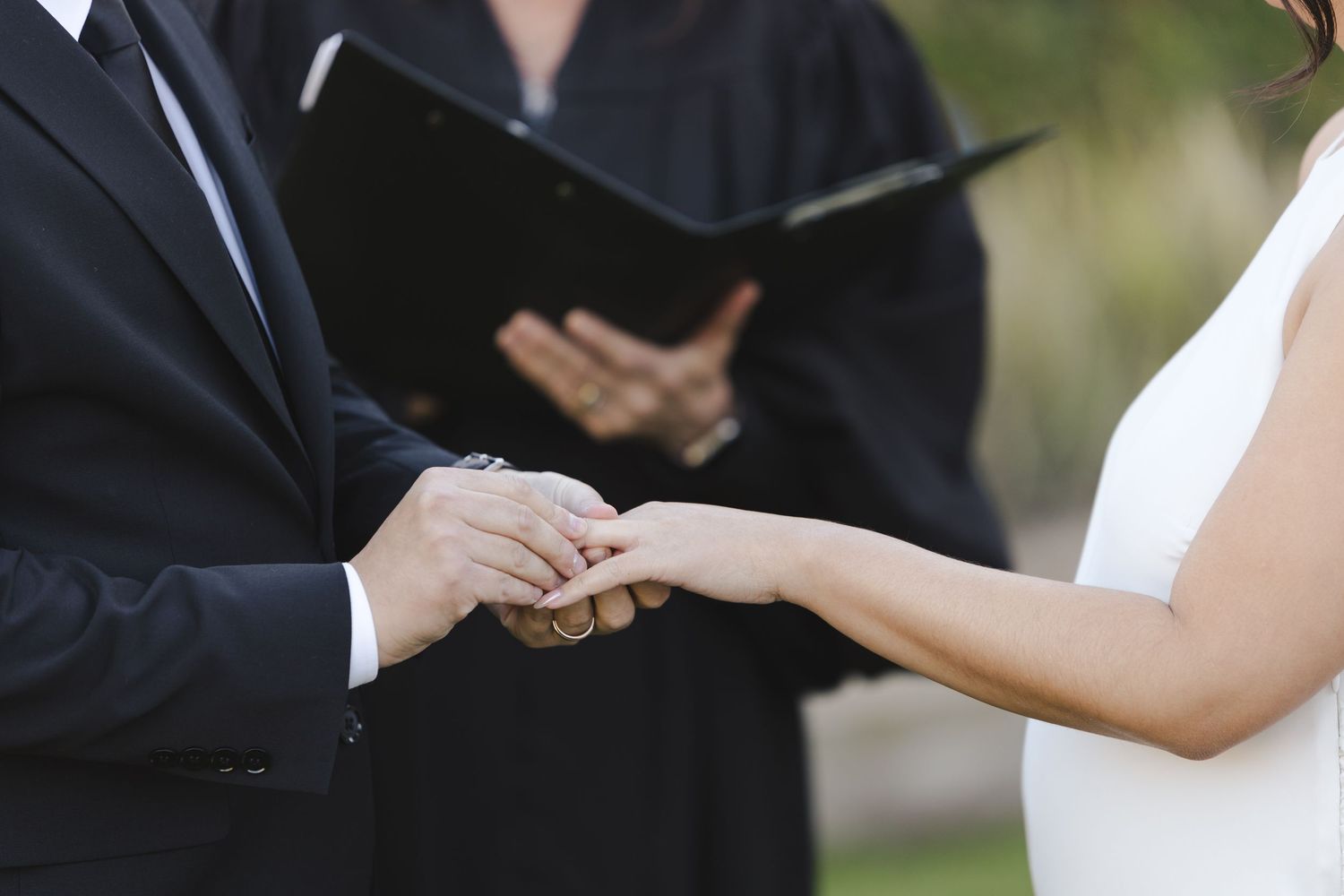A couple exchanges rings during their wedding ceremony with an officiant in a dark robe standing behind them.