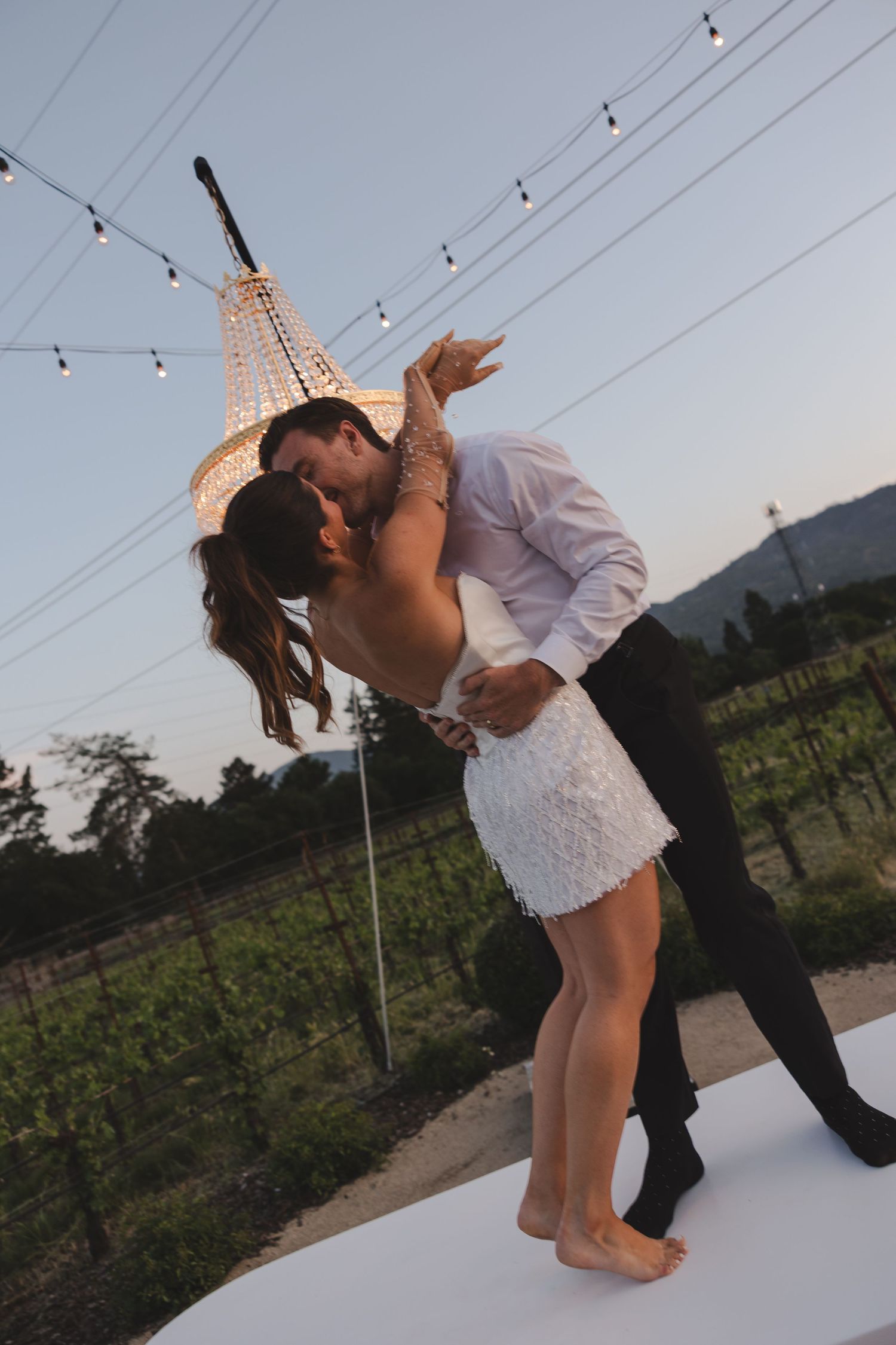 A romantic dip dance captured outdoors with string lights overhead and mountains in the background at sunset.