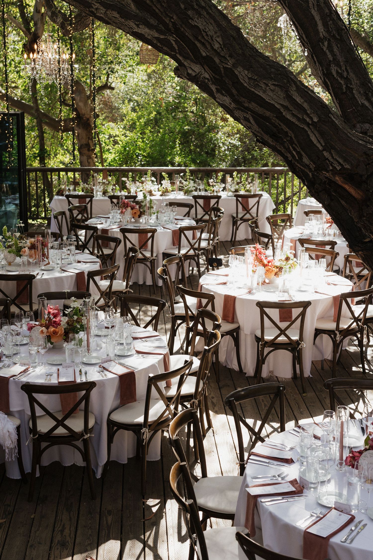 Outdoor wedding reception with round tables, cross-back chairs, and floral centerpieces under trees.