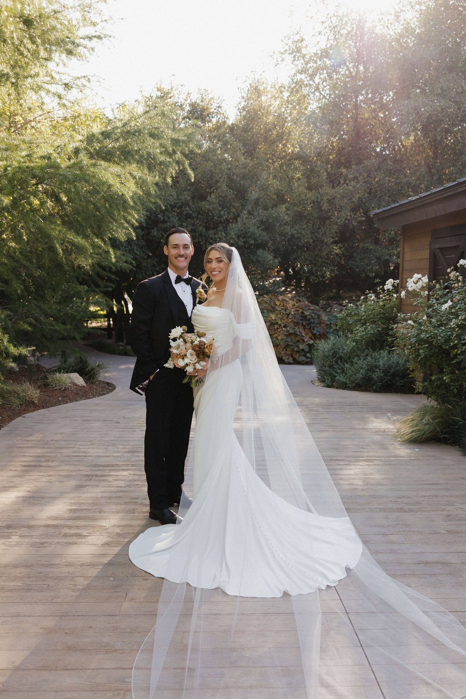 A wedding couple poses on a stone patio surrounded by lush greenery at golden hour.