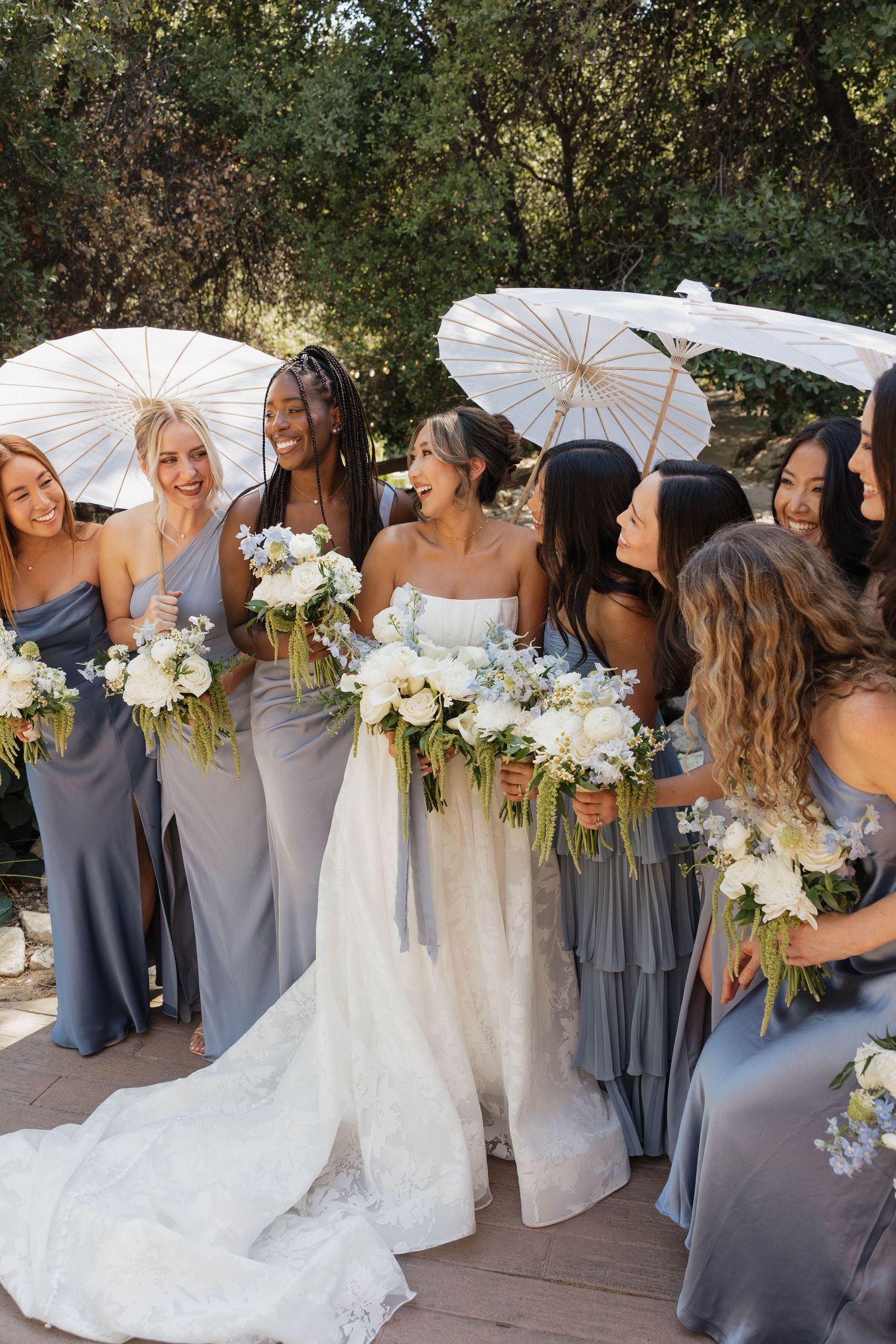Wedding party members holding white parasols and floral bouquets pose for photos in an outdoor garden setting.