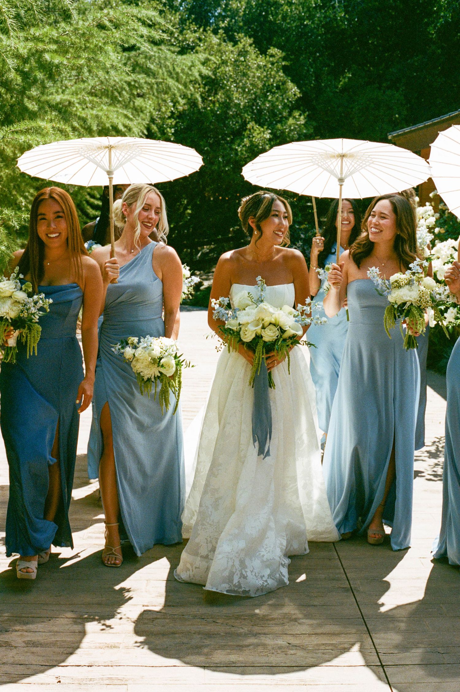 Wedding party with bridesmaids in light blue dresses holding white parasols alongside bride with bouquet.