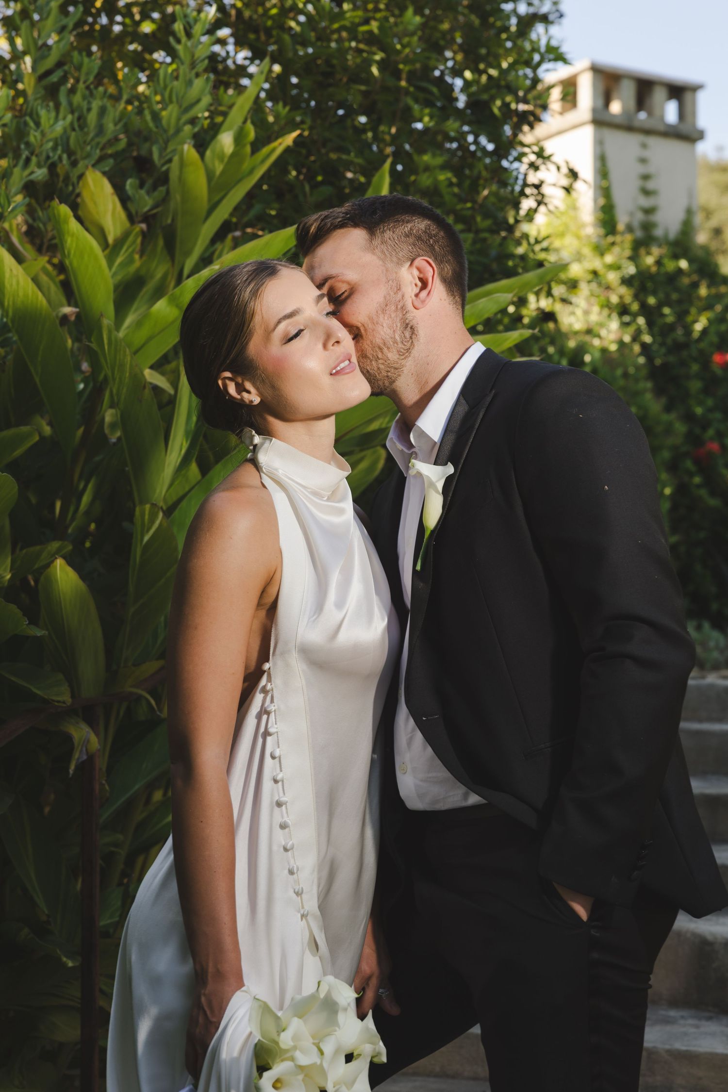 A romantic wedding couple share a tender moment in a garden setting with a white stone tower in the background.