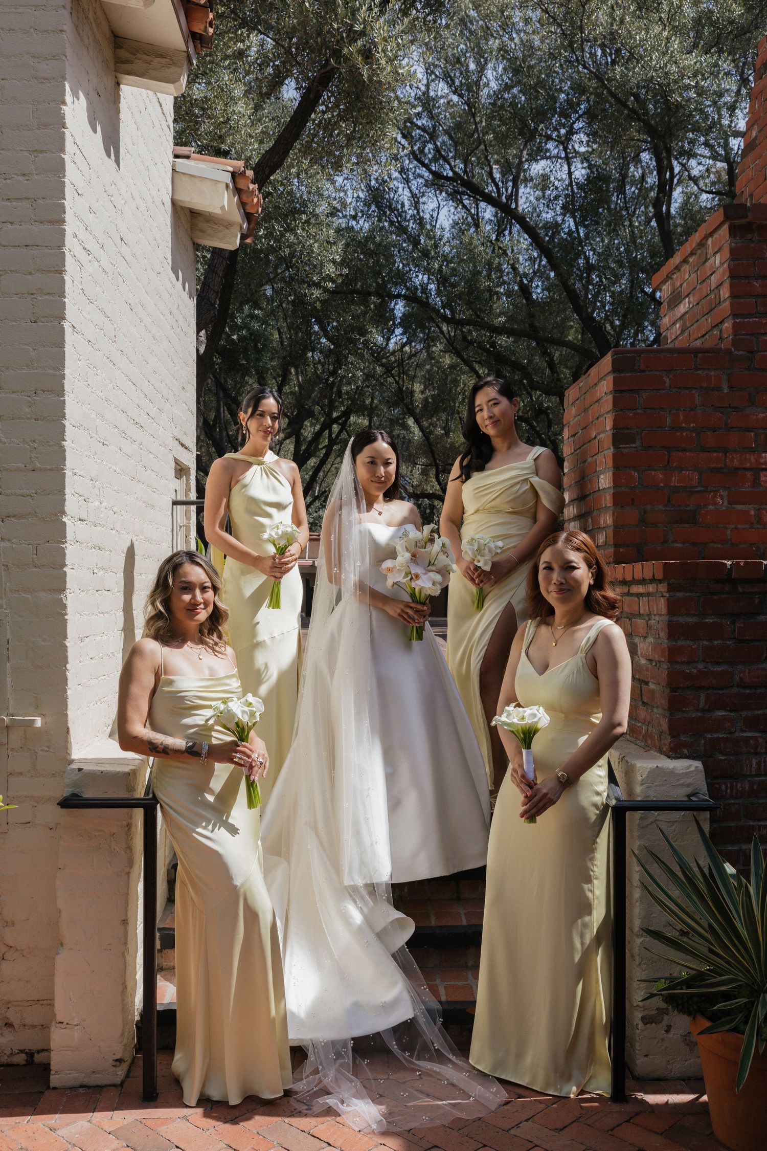 Bride in white gown with four bridesmaids in sage green dresses holding bouquets on sunlit outdoor steps.