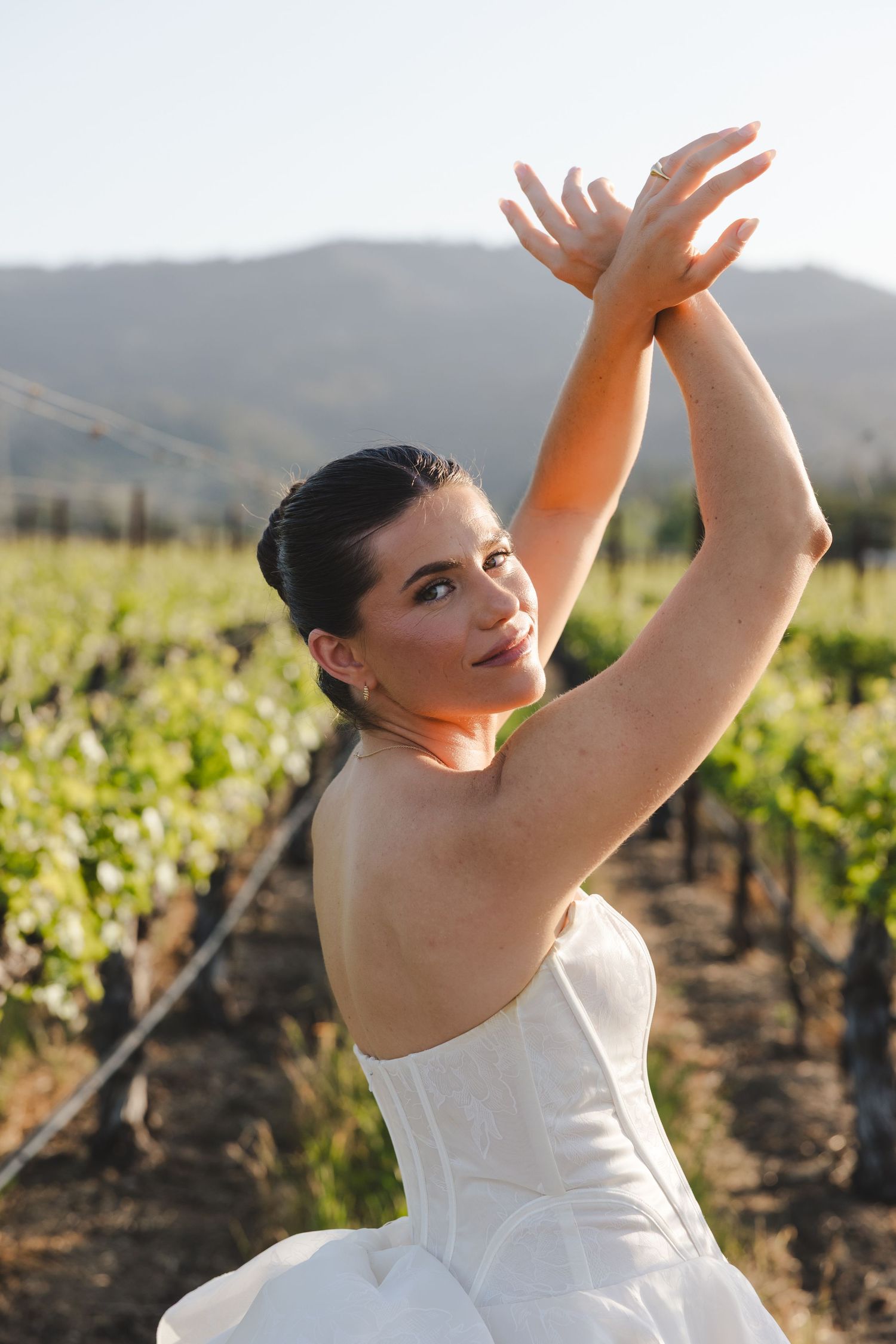 A graceful figure in a white strapless gown poses with raised arms in a sunlit vineyard.