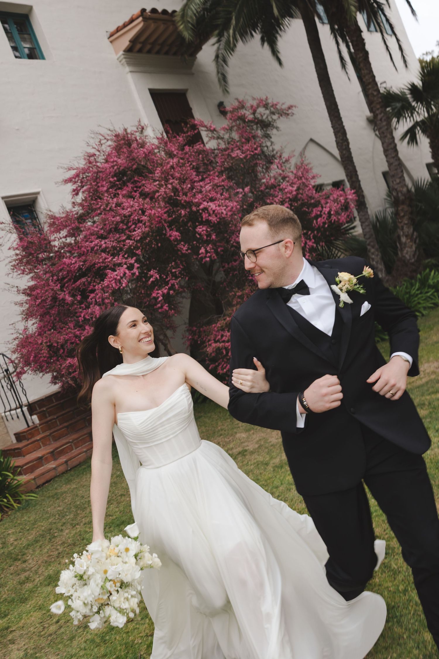 A couple in formal wedding attire dance and laugh together in front of a white building with pink bougainvillea flowers.