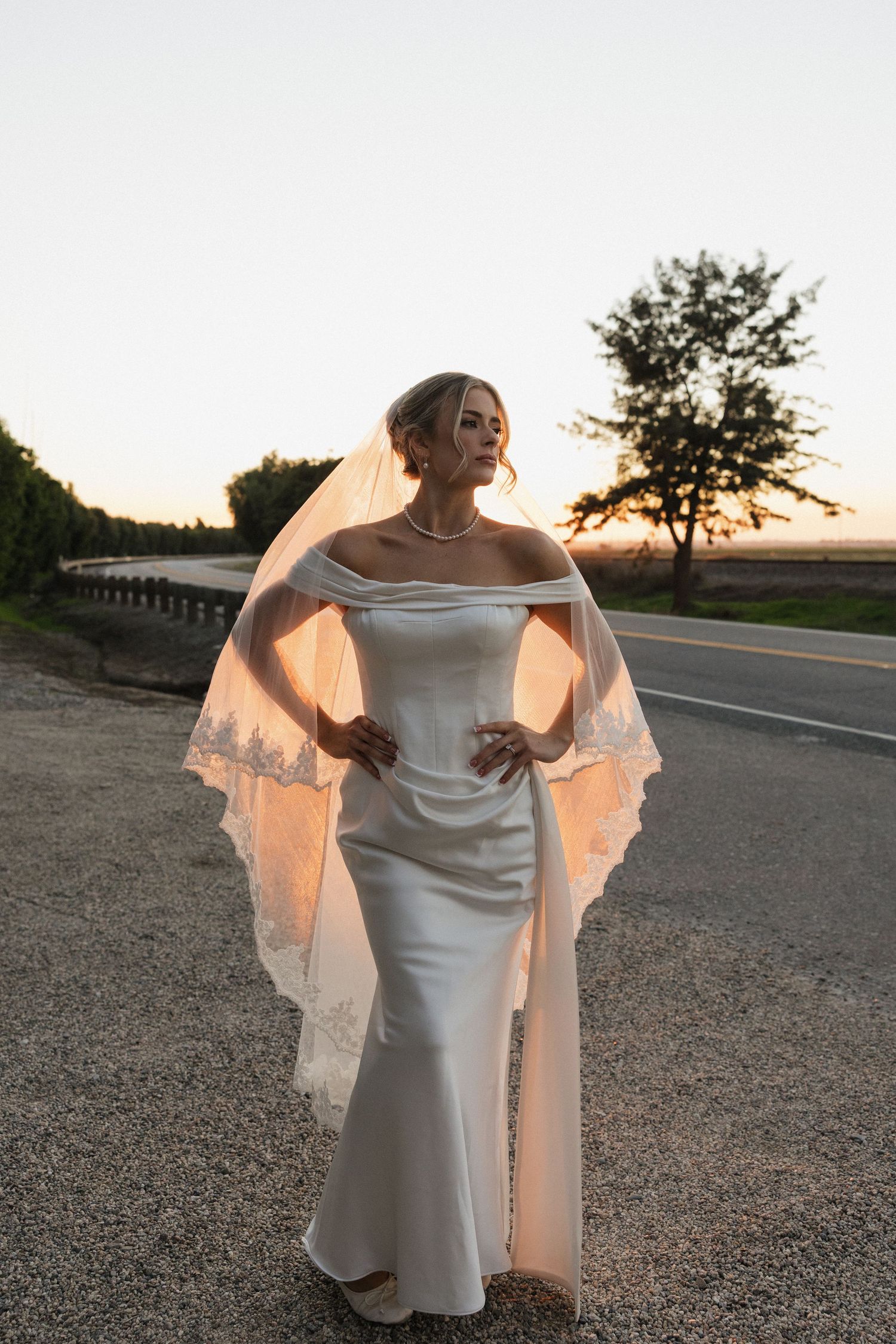 Elegant bride in off-shoulder white dress poses at sunset on a gravel path.