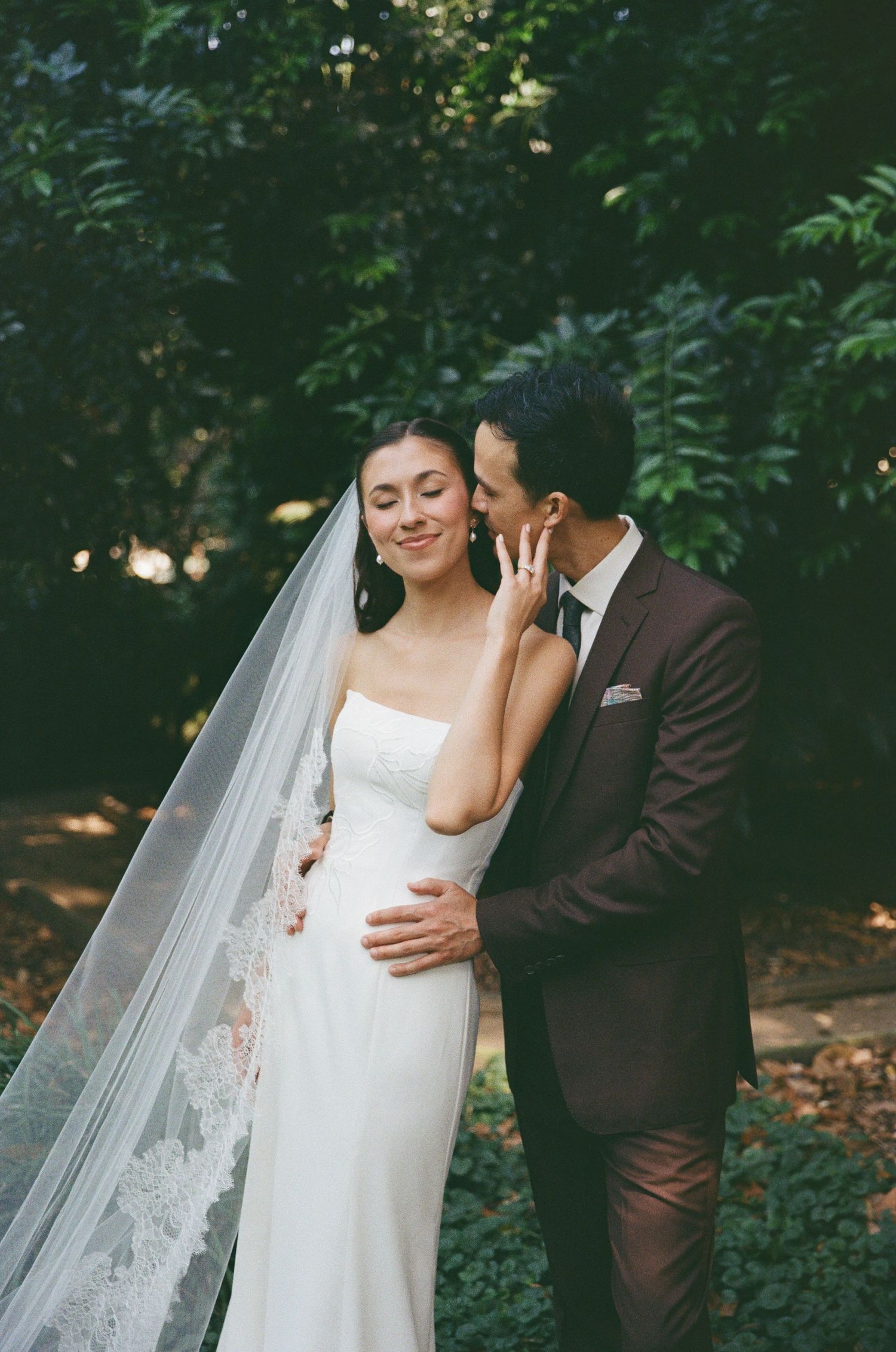 A bride in a white gown and lace veil smiles as her groom in a brown suit whispers in her ear outdoors.